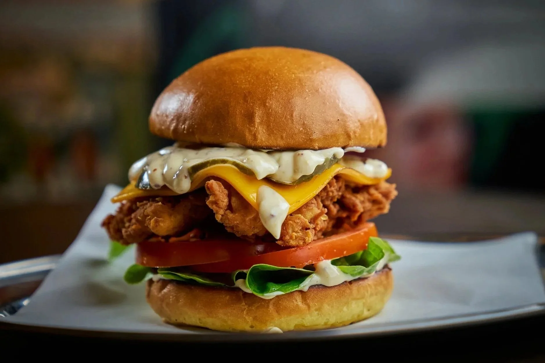 Close-up of a multi-layered fried chicken sandwich with lettuce, tomato, cheese, pickles, mayonnaise, and a toasted bun, on a white plate.