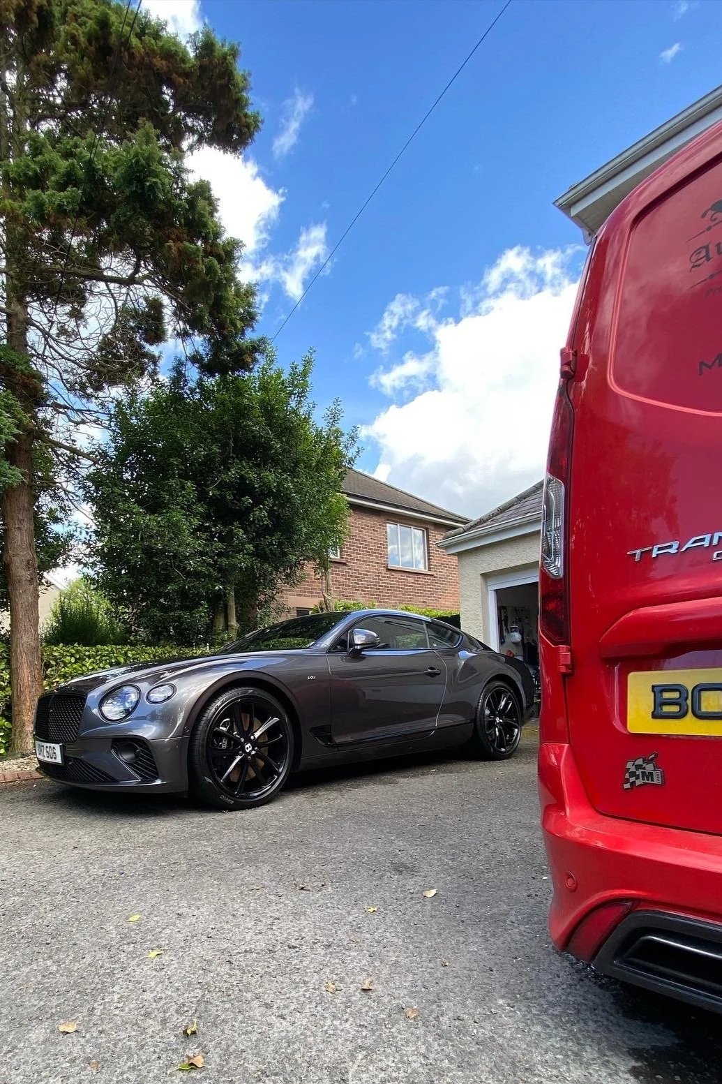 There is a gray sports car parked in a driveway with a red van partially blocking the view. The background shows a brick house, trees, and a blue sky with some clouds.