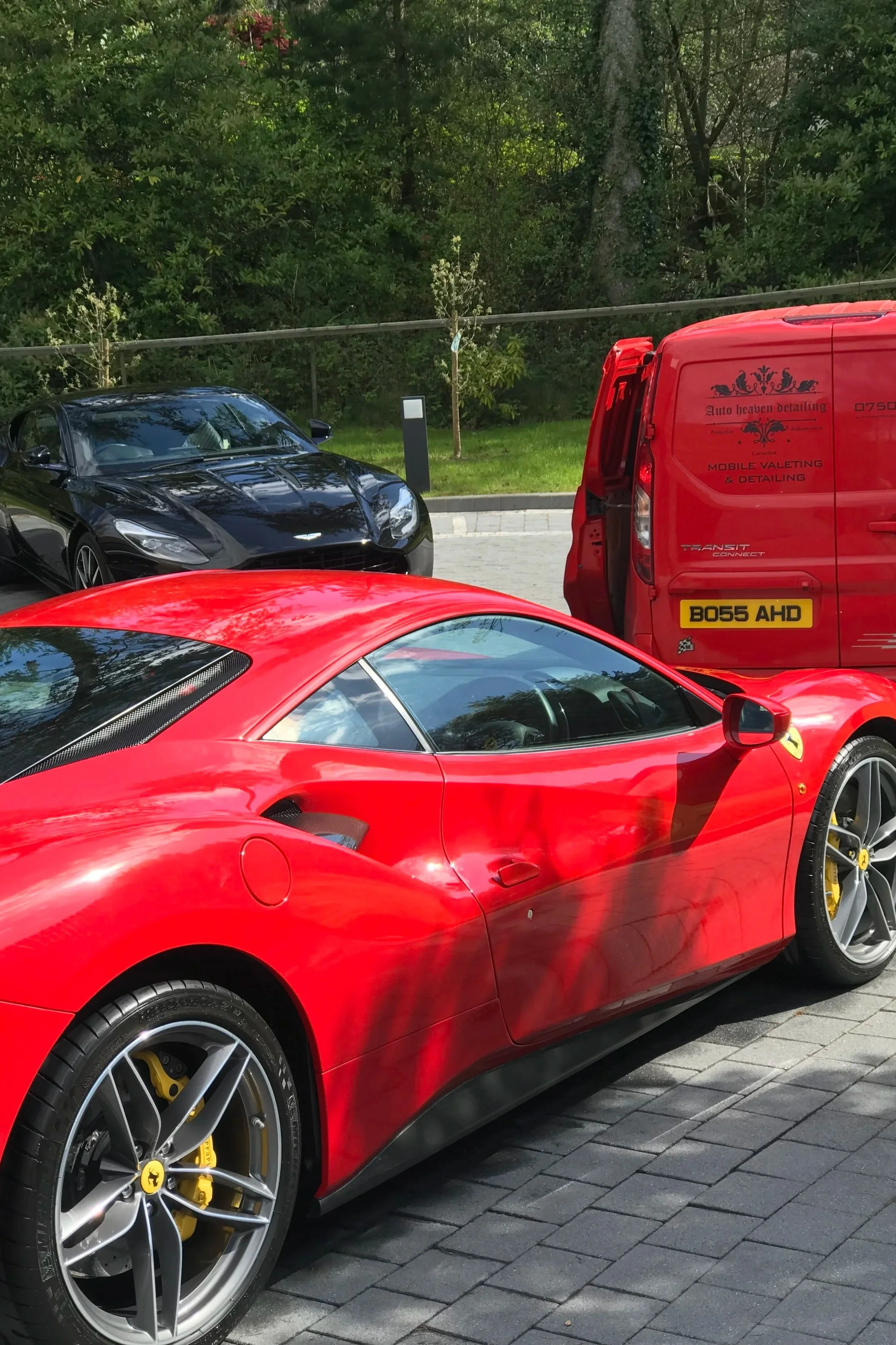 Red Ferrari sports car parked on a street with a black Aston Martin and a red van nearby, surrounded by green trees and a paved sidewalk.
