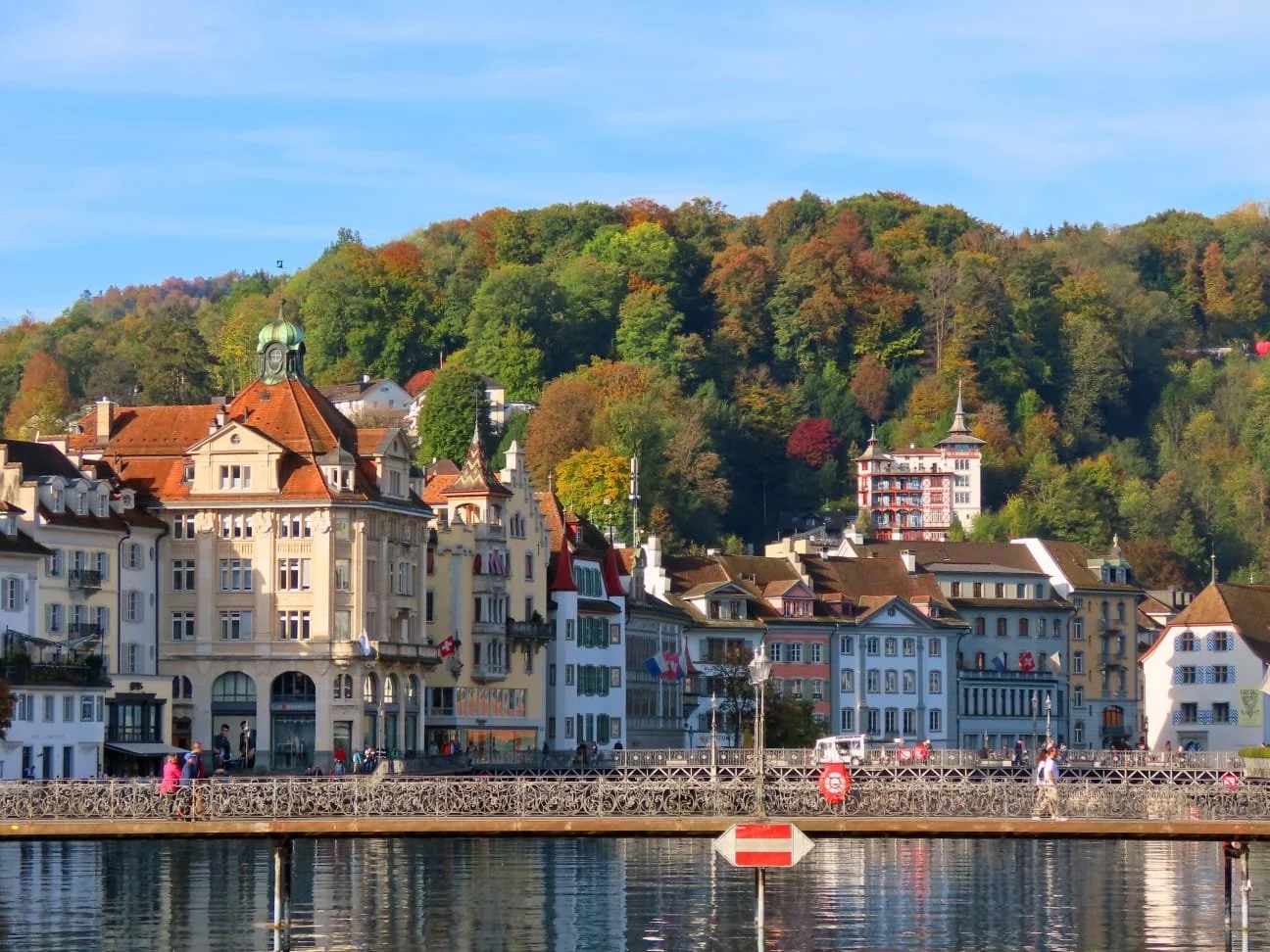 Scenic view of a European lakeside town with colorful historic buildings, a wooden bridge with railings, and a green hill with autumn trees in the background under a blue sky.