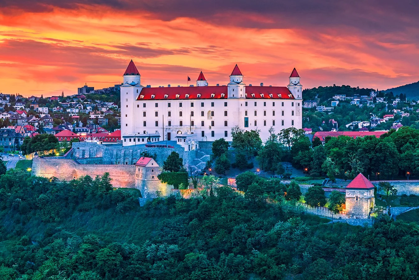 A large white castle with red roofs and four towers is situated on a hill, surrounded by lush green trees and a small town at sunset.