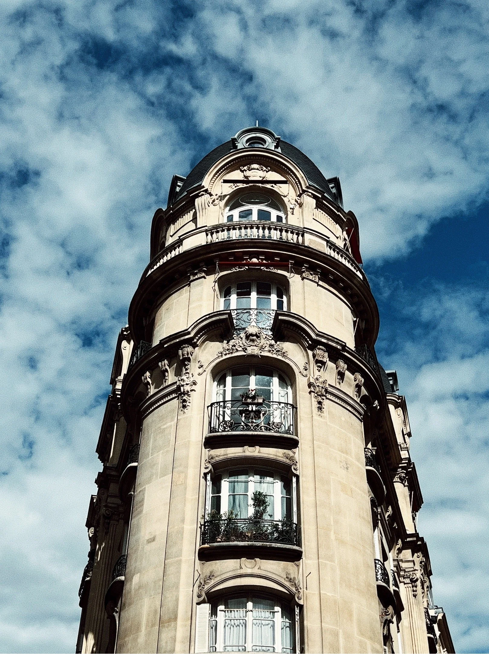 A historic multi-story building with rounded windows, intricate architectural details, and decorative balconies, set against a cloudy blue sky.