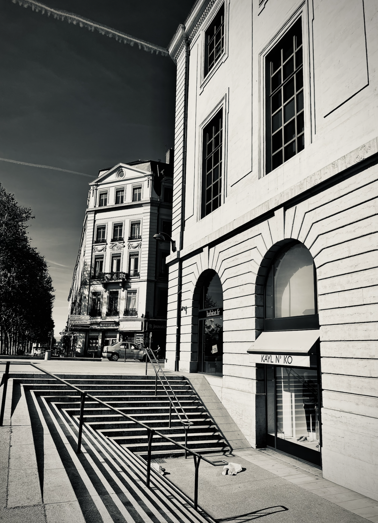 Black and white photo of classical European-style building with arched windows and a staircase with metal railings in the foreground. A small sign on the building reads 'KAYL N'KO'.