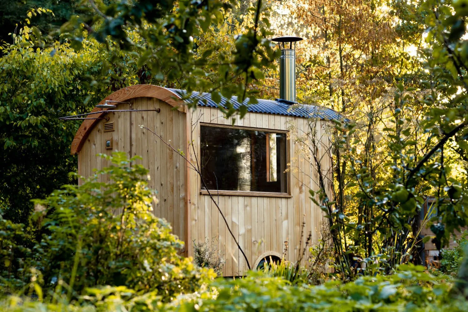 Wood sided sauna with an outdoor shower on one end, a large window on the side facing us and a tall galvanised metal chimney on top