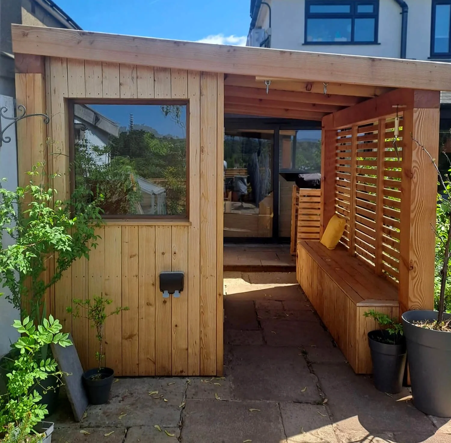 Small sauna in a back garden with a window and a wooden slatted screen and a storage bench
