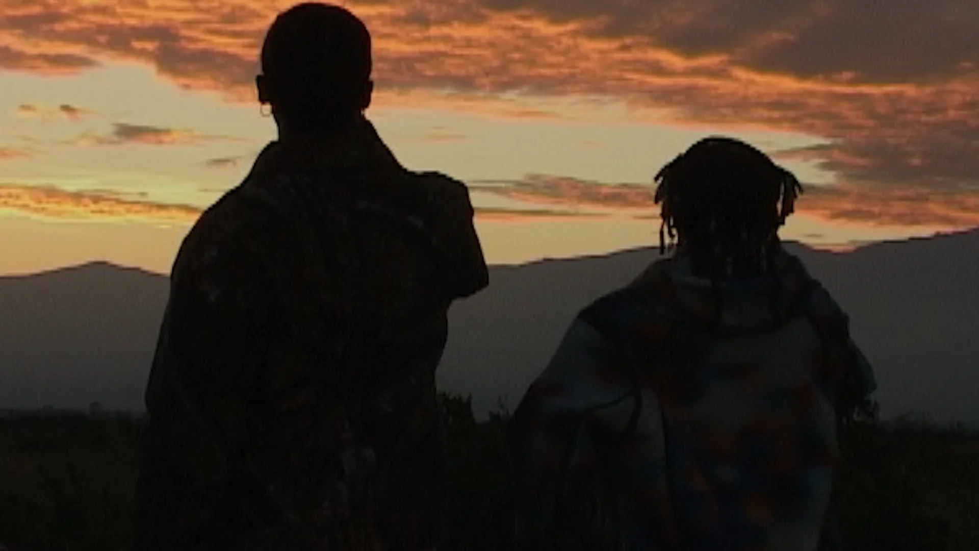 Two people, one with short hair and one with dreadlocks, standing outdoors during sunset with their backs to the camera, overlooking distant mountains and a colorful sky.