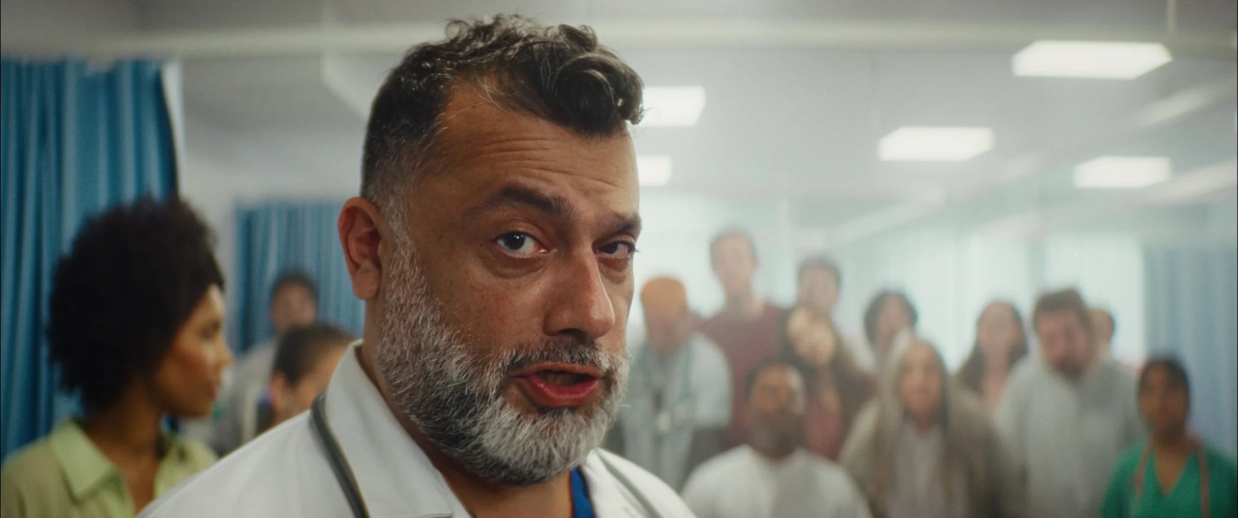A close-up of a male doctor with gray hair and a beard, wearing a white coat with a stethoscope, speaking in a hospital corridor with a group of diverse people in the background.