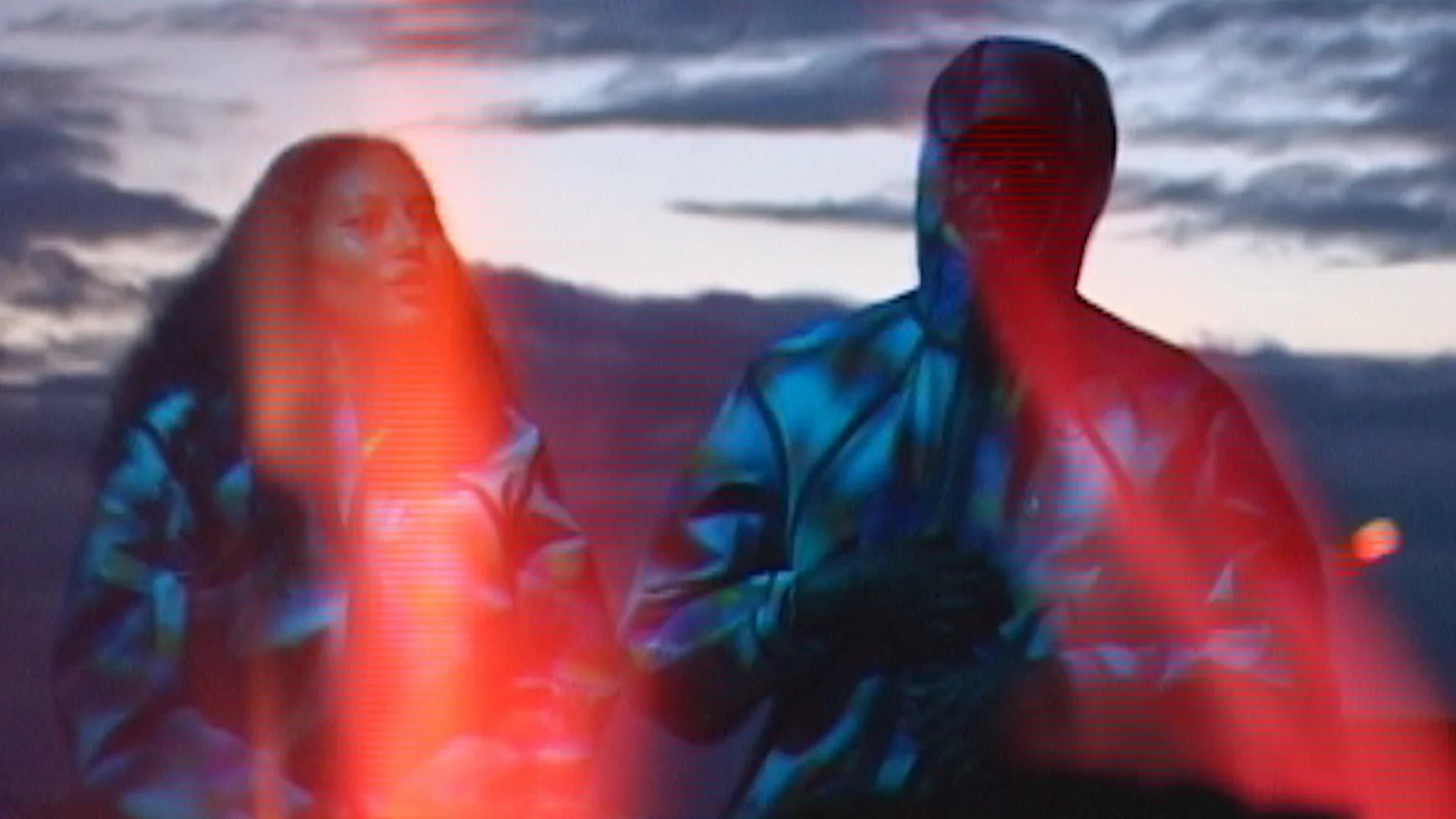 Two people sitting outdoors during sunset, wearing colorful clothing, with cloudy sky in the background.