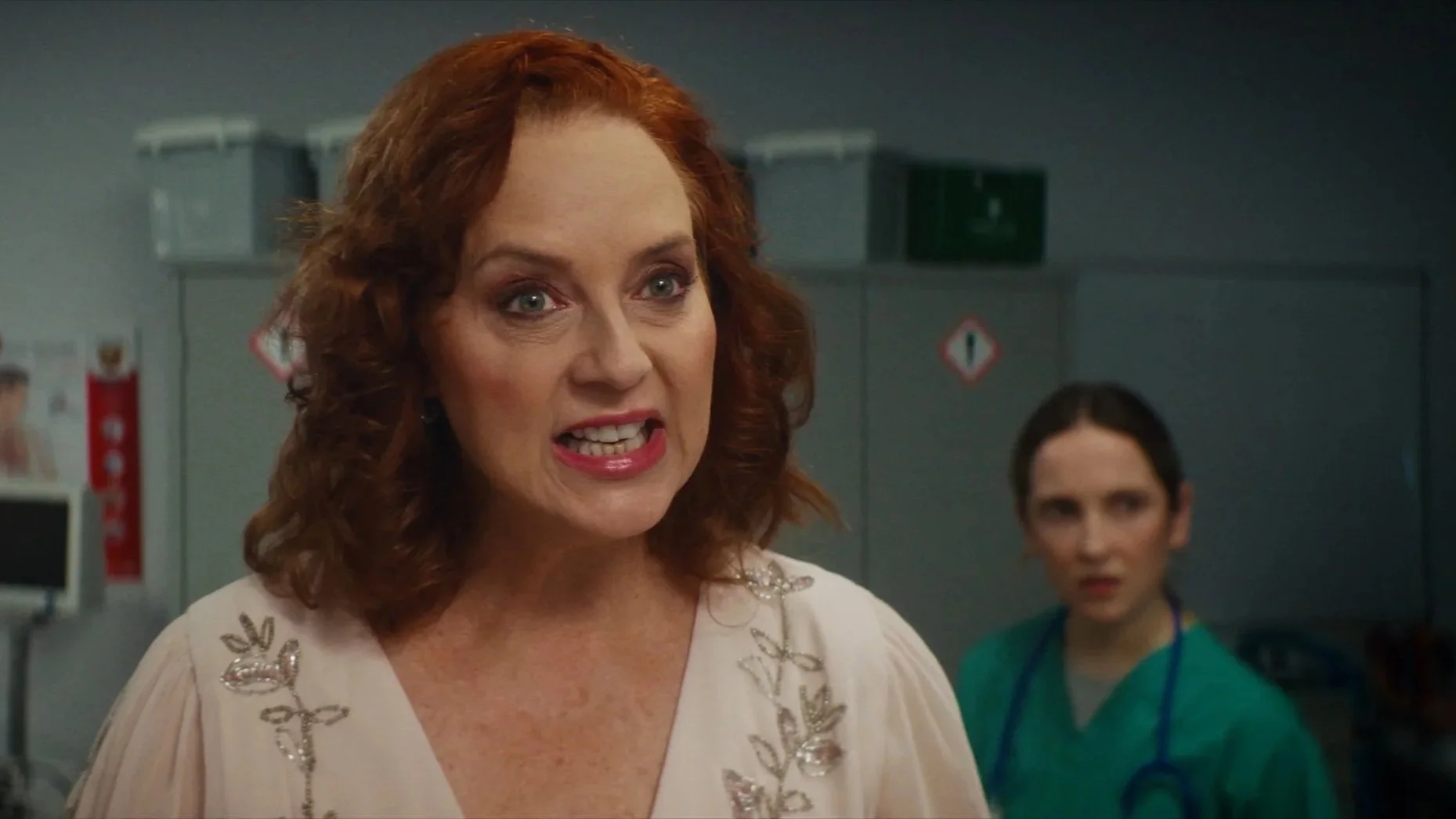 A woman with red curly hair speaking, wearing a cream-colored dress with embroidered leaf designs, in a medical setting with a young female healthcare worker in scrubs in the background.