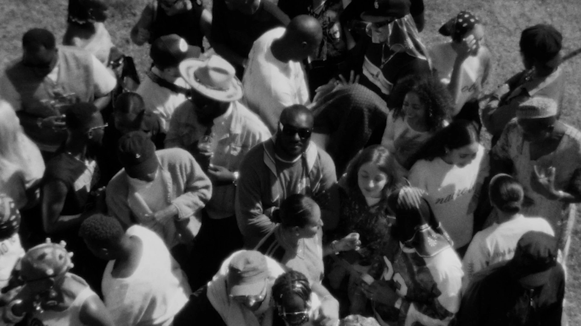 Crowd of diverse people, including men and women, gathered outdoors at night, some wearing hats and sunglasses, engaged in conversation and socializing.