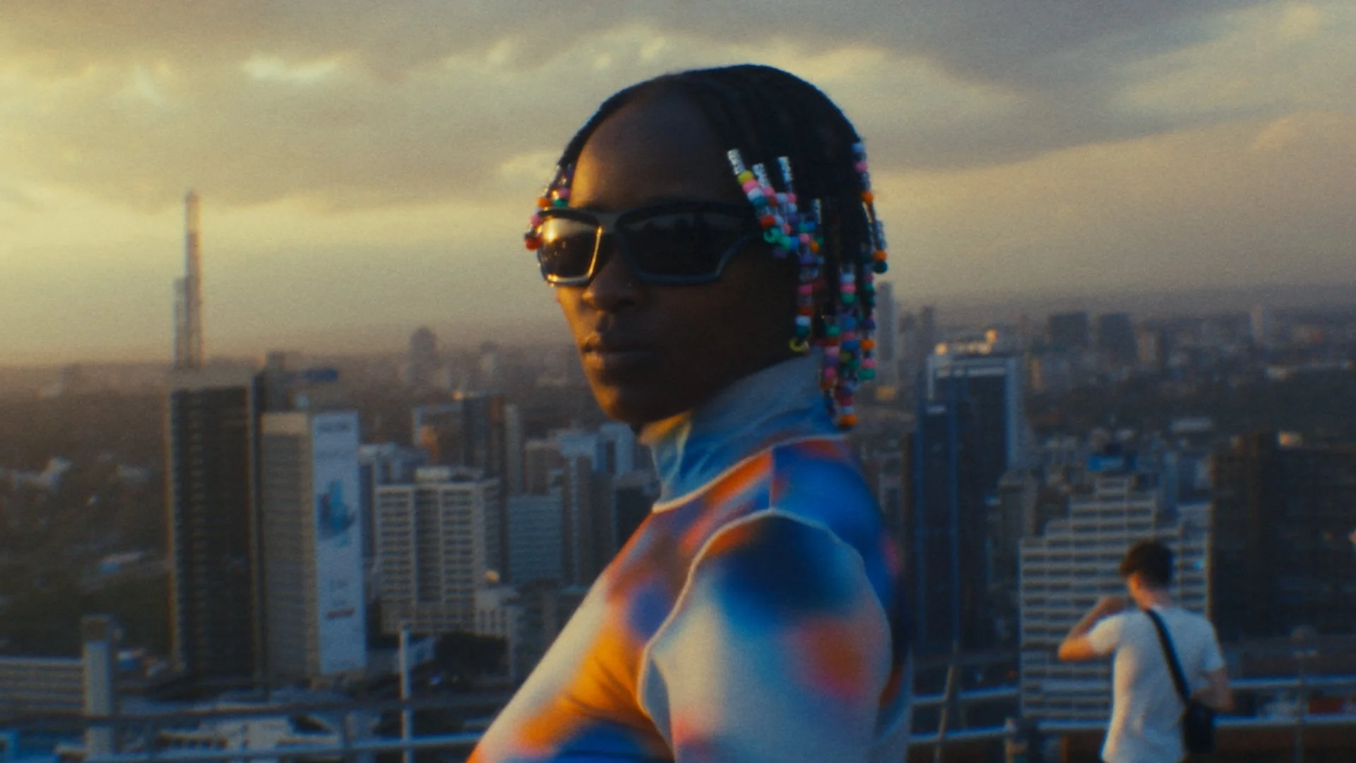 Person with colorful beaded hair and sunglasses looking over city skyline at sunset.