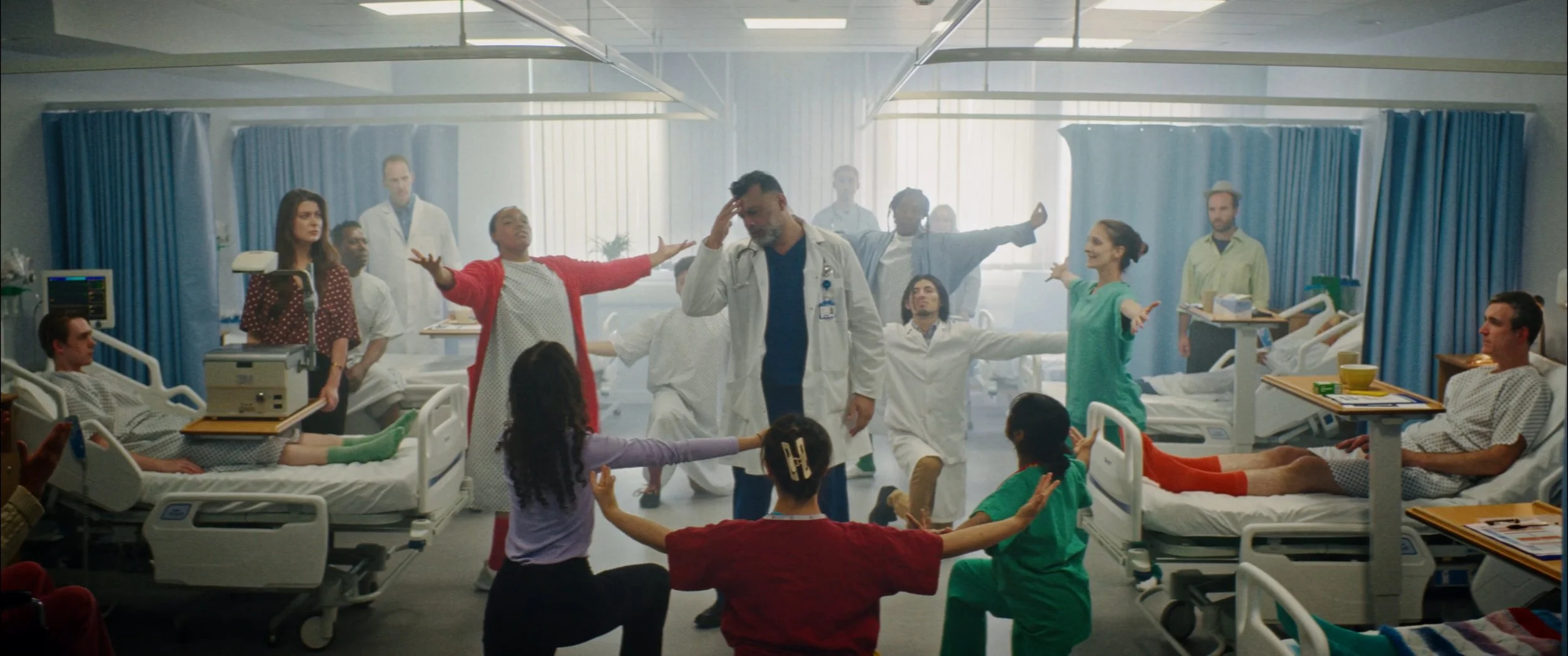 Hospital scene with patients in beds and healthcare workers standing and sitting, some with arms outstretched, in a bright, modern ward.