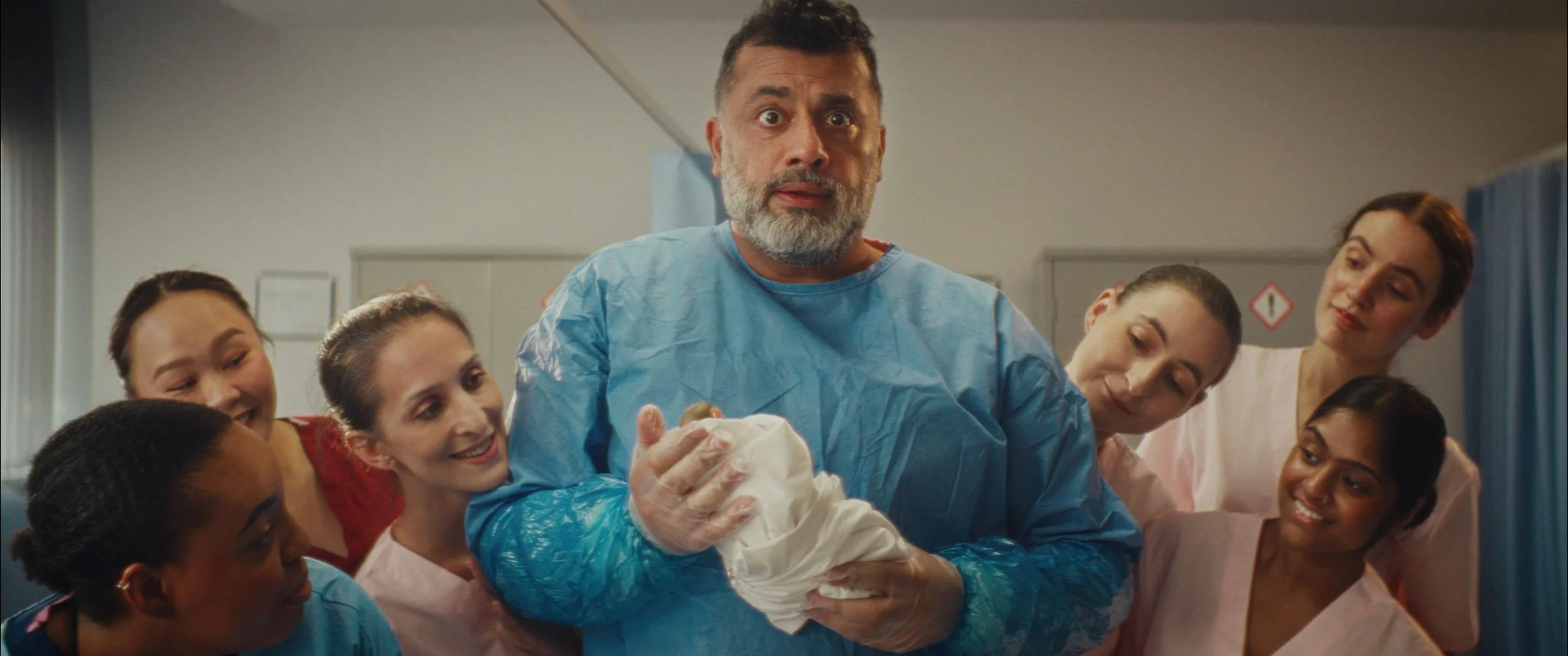 Man in medical gown holding newborn baby, surrounded by smiling healthcare workers in a hospital corridor.