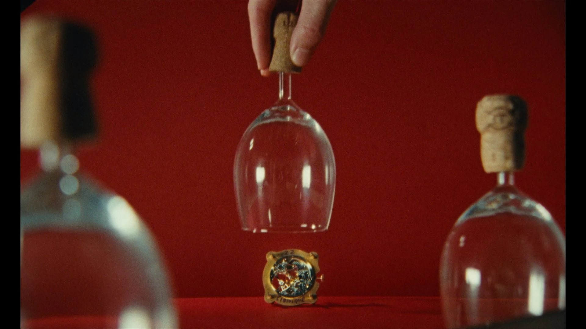 Multiple upside-down wine glasses with cork stoppers hanging in a row against a red background, with a charm or decorative item with a colorful design below.