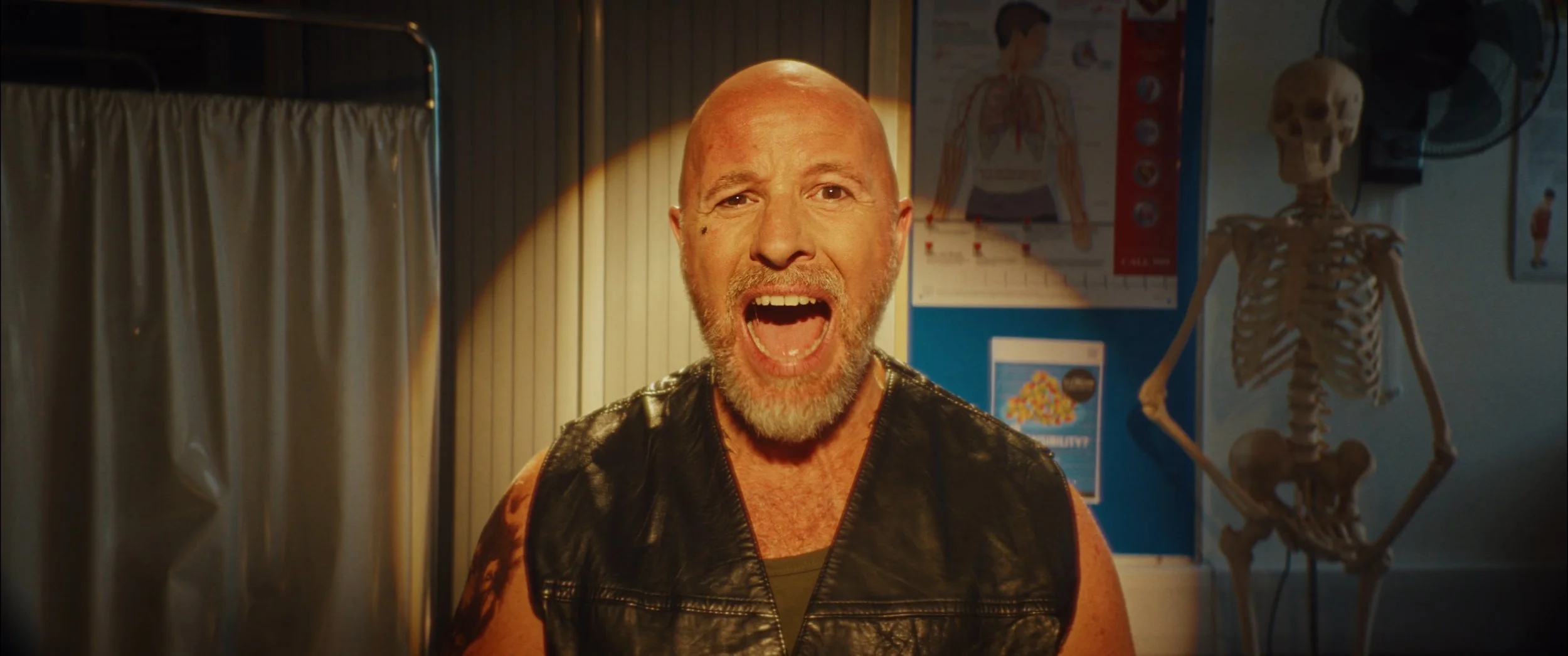 A man with a bald head and a beard, wearing a black leather vest, screaming with an open mouth inside a room with medical posters and a skeleton model in the background.