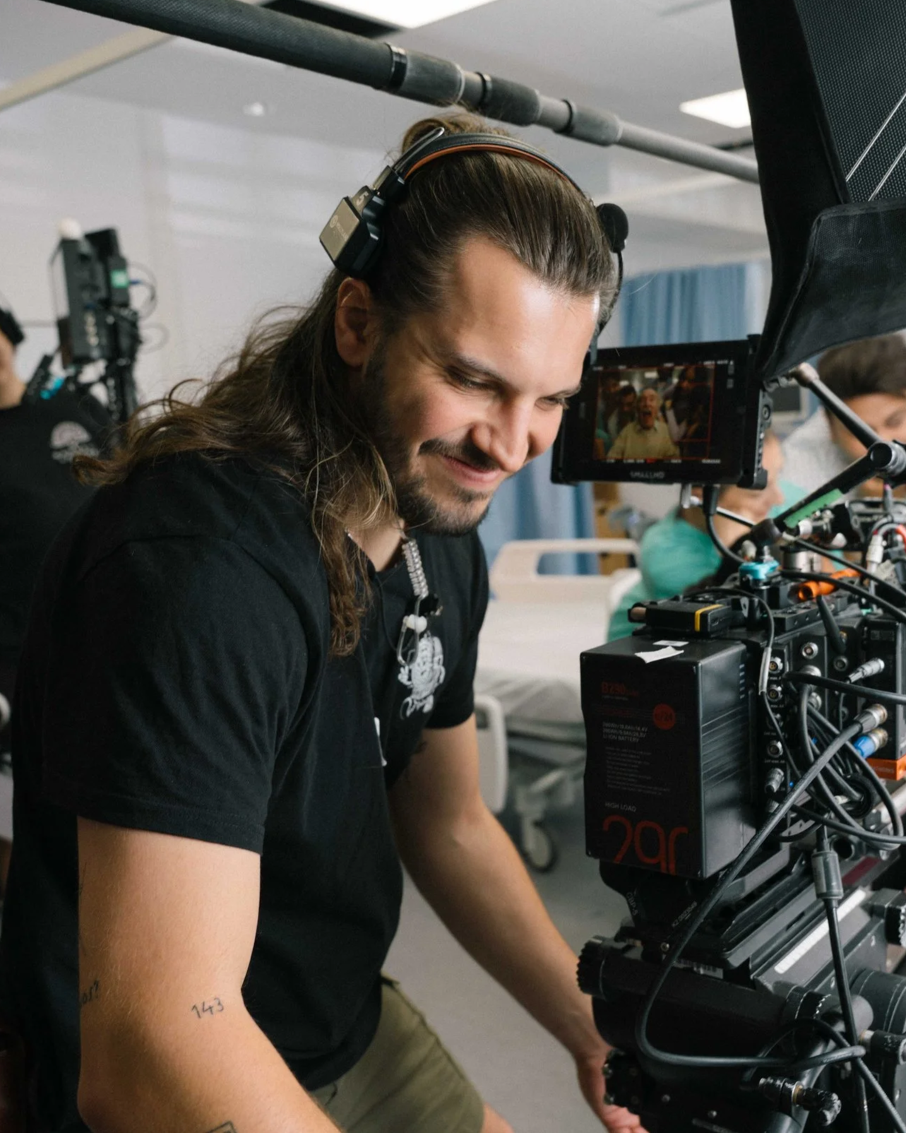 A man with long hair and a beard, wearing headphones, operates a professional camera on a film set, with a blurred background of other people and equipment.