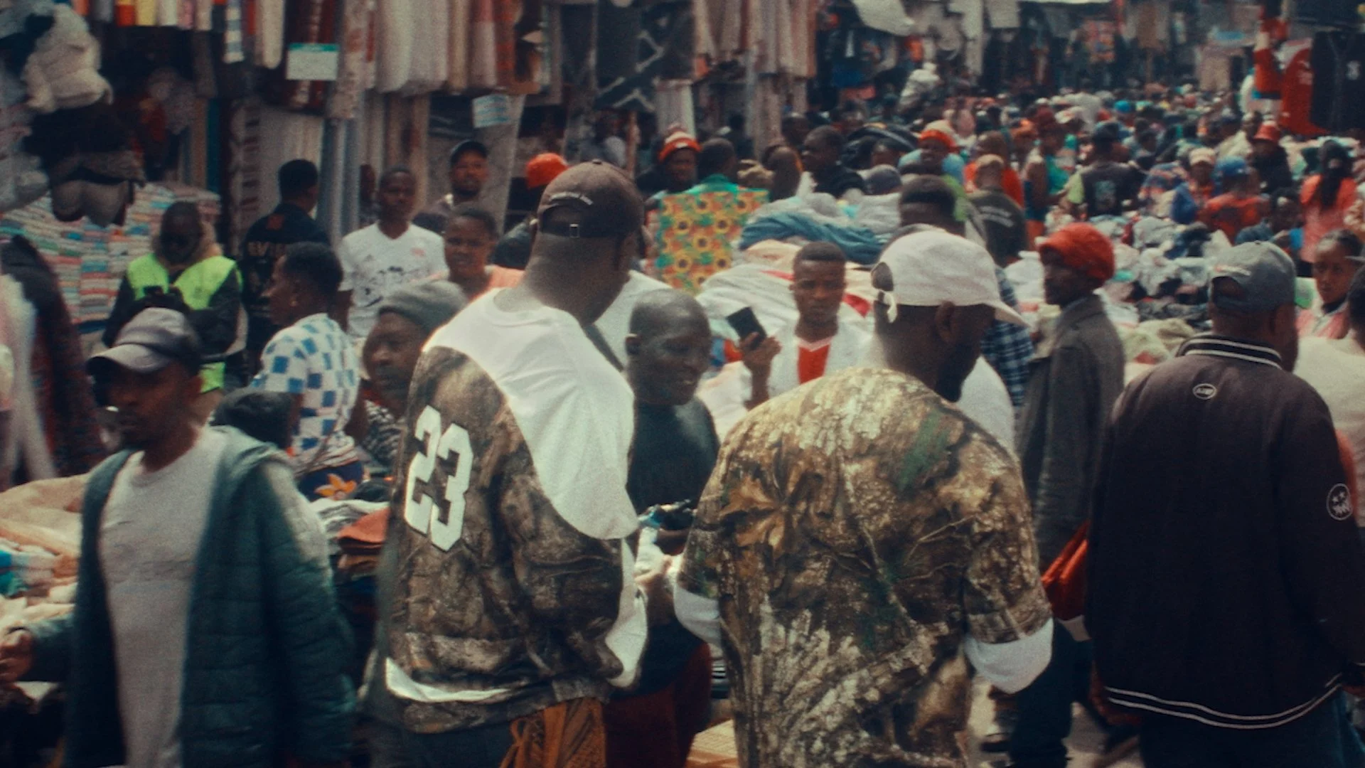 Crowded market street with numerous people shopping, some holding phones, women and men wearing caps and colorful clothing, shops on both sides selling clothing and fabrics.