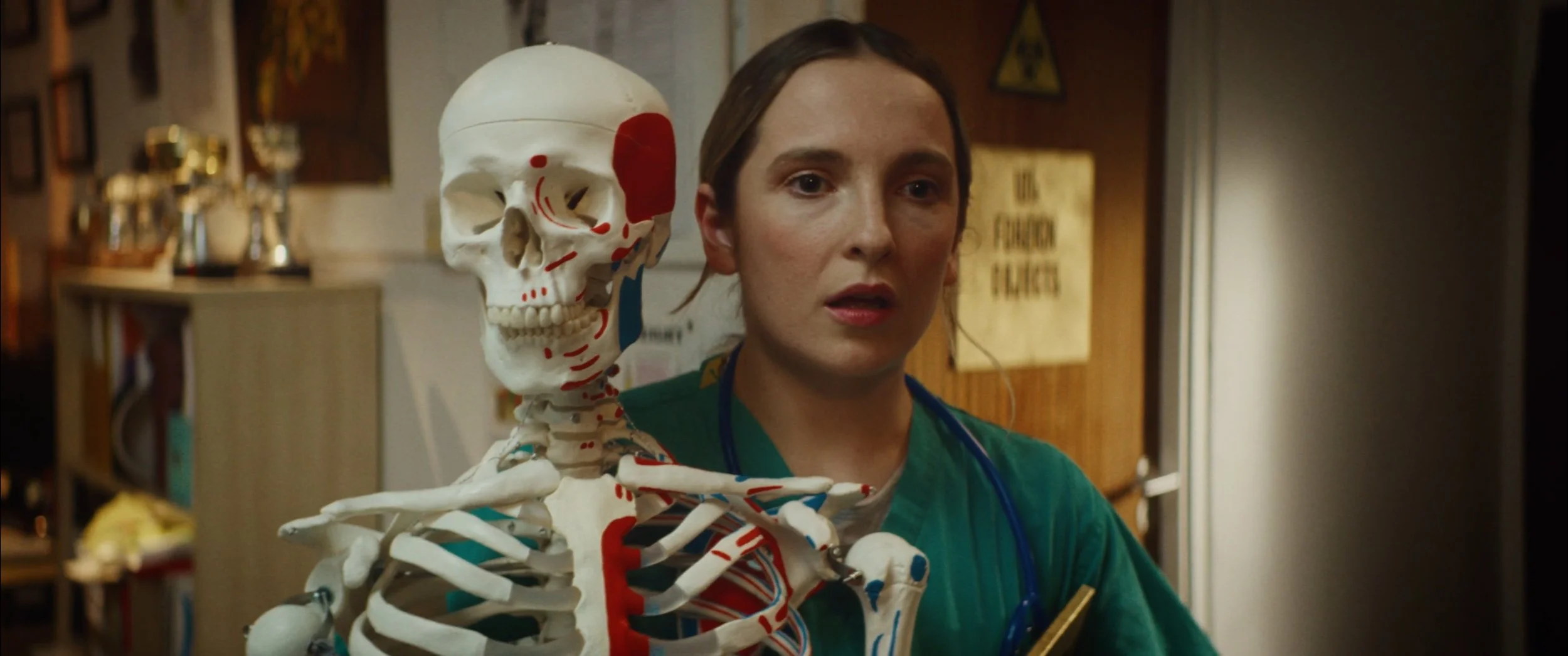 A woman in medical scrubs with a stethoscope around her neck standing next to a life-sized anatomical skeleton model in a medical setting.