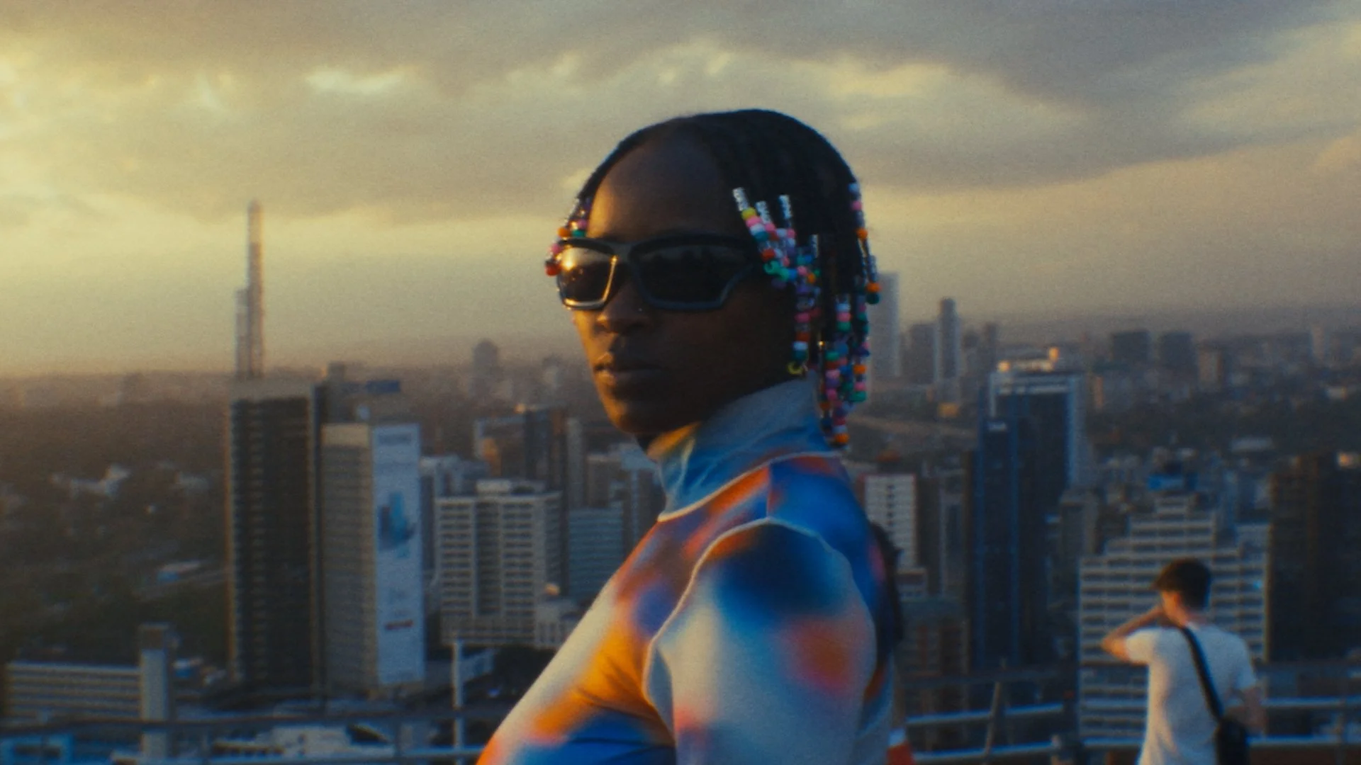 A person with braided hair decorated with colorful beads wears dark sunglasses and a vibrant jacket, standing on a rooftop overlooking a cityscape at sunset.