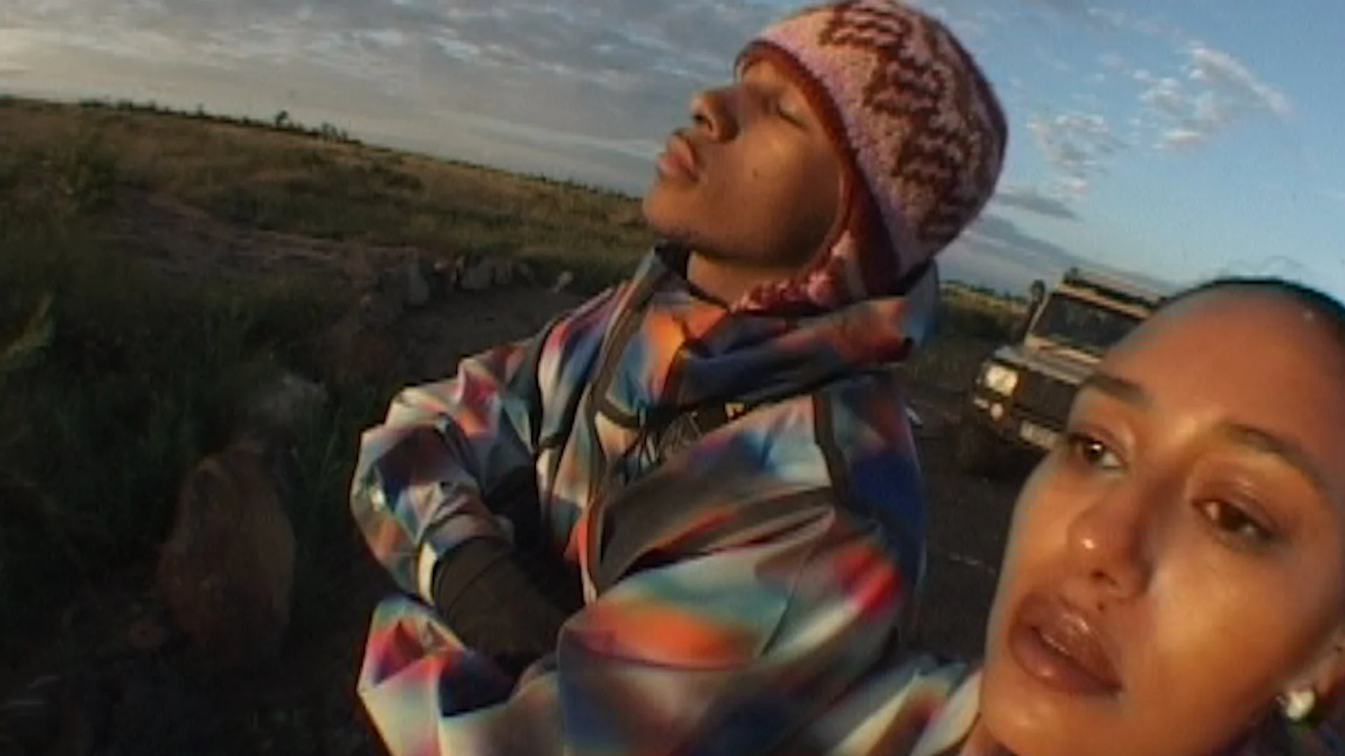 Two women in colorful jackets and a woman in a multicolored beanie are outdoors, with a field and a truck in the background. One woman has her eyes closed and the other is smiling.