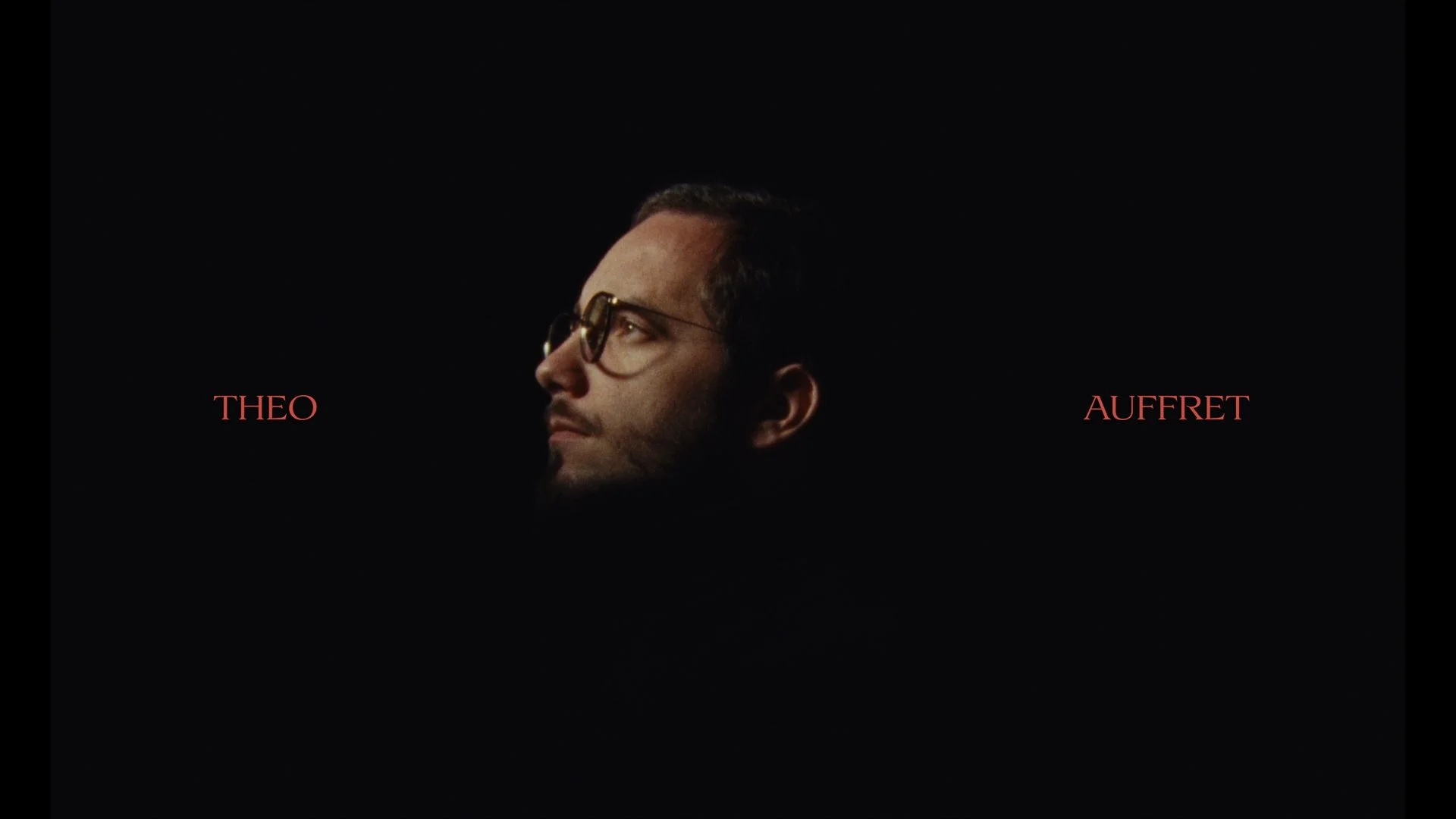 Close-up of a man with glasses looking to the left, surrounded by black space, with the words 'THEO' on the left and 'AUFFRET' on the right in red text.