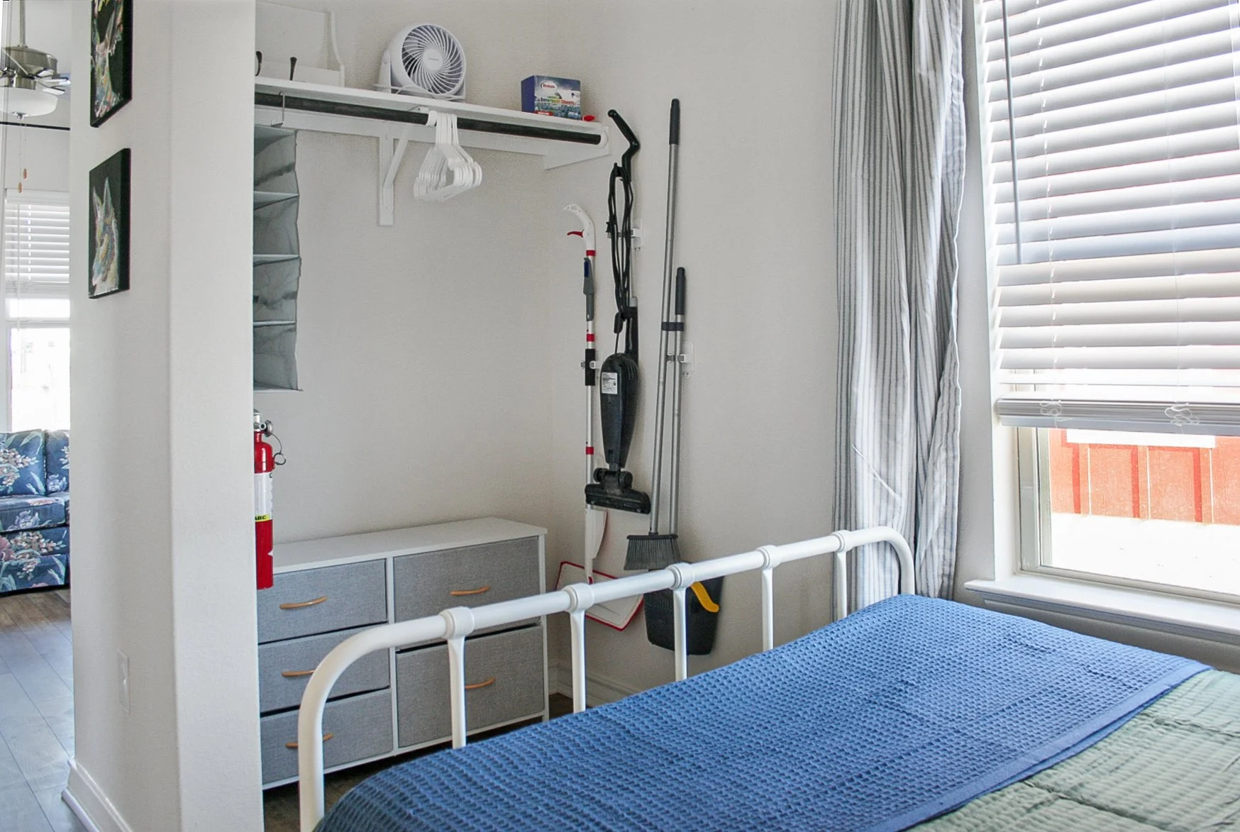 A bedroom with a white metal bed frame, a blue blanket on the bed, a dresser, and a closet area with cleaning tools and a small fan. There is a window with blinds and curtains letting in natural light.