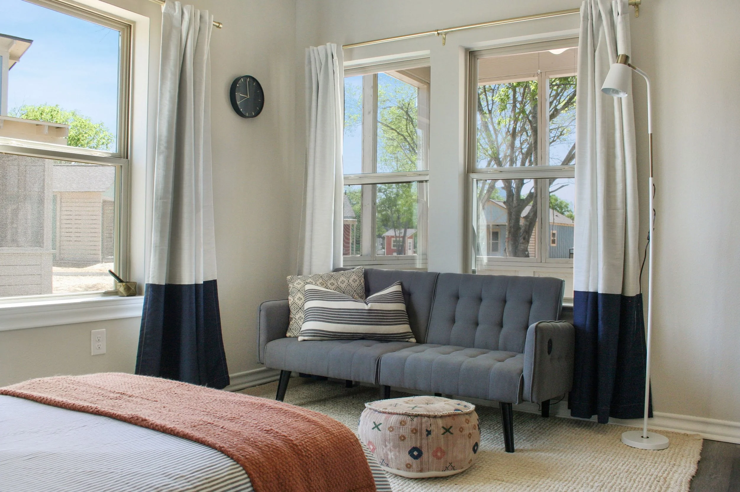 A cozy living room corner with a gray sofa, decorative pillows, a round patterned ottoman, a floor lamp, and three windows with white curtains allowing natural light.