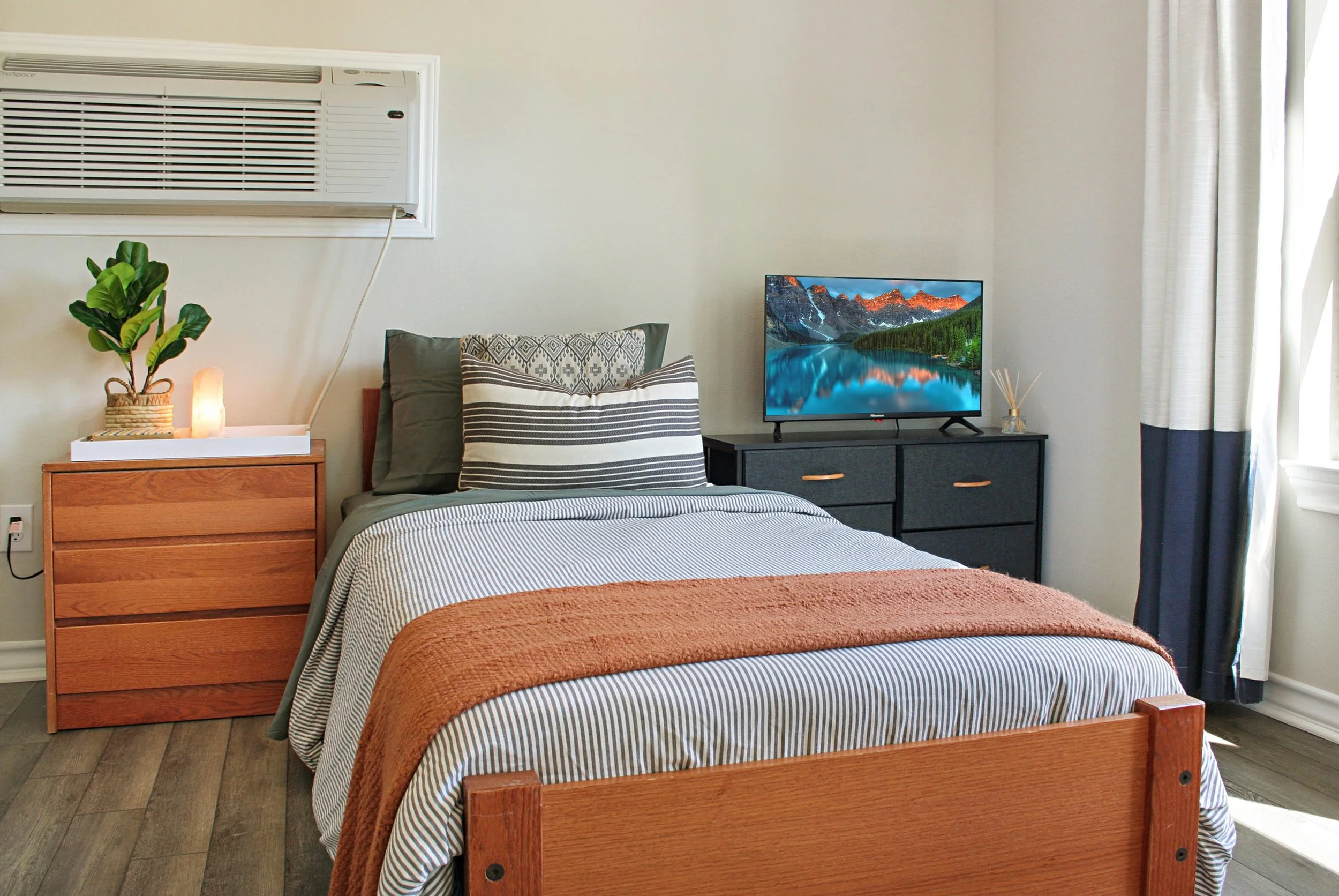 A bedroom with a wooden bed, striped bedding, and a large throw blanket. A nightstand with a plant, salt lamp, and an air conditioning unit above. A dark dresser with a flat-screen TV, a reed diffuser, and decor near a window with curtains.