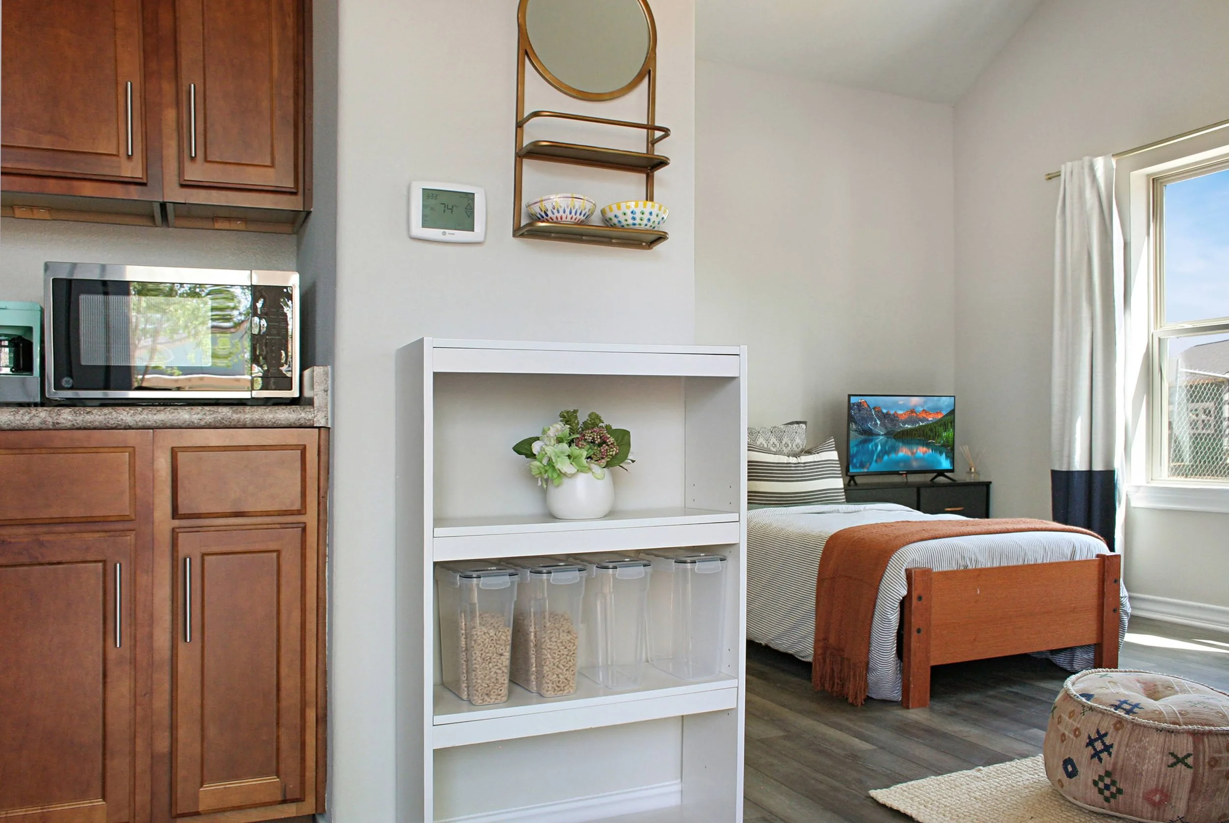 A small kitchen area with brown cabinets, a microwave, a white shelf with containers and a flower vase, and a bedroom with a bed, window, and a TV in the background.