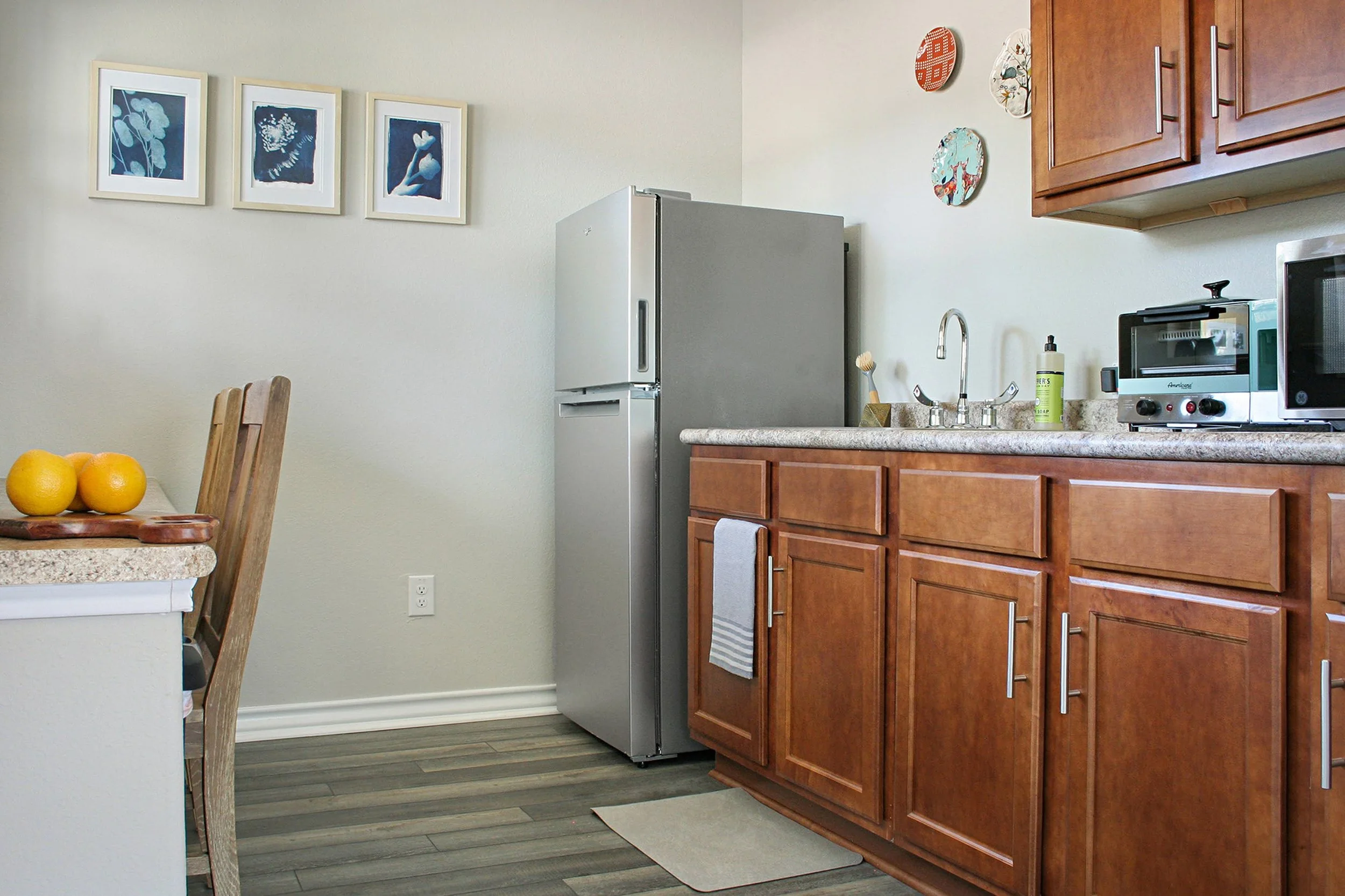 Kitchen with wooden cabinets, a silver refrigerator, a sink with a soap dispenser, and small appliances like a toaster oven and microwave. There are decorative plates on the wall and a table with lemons.