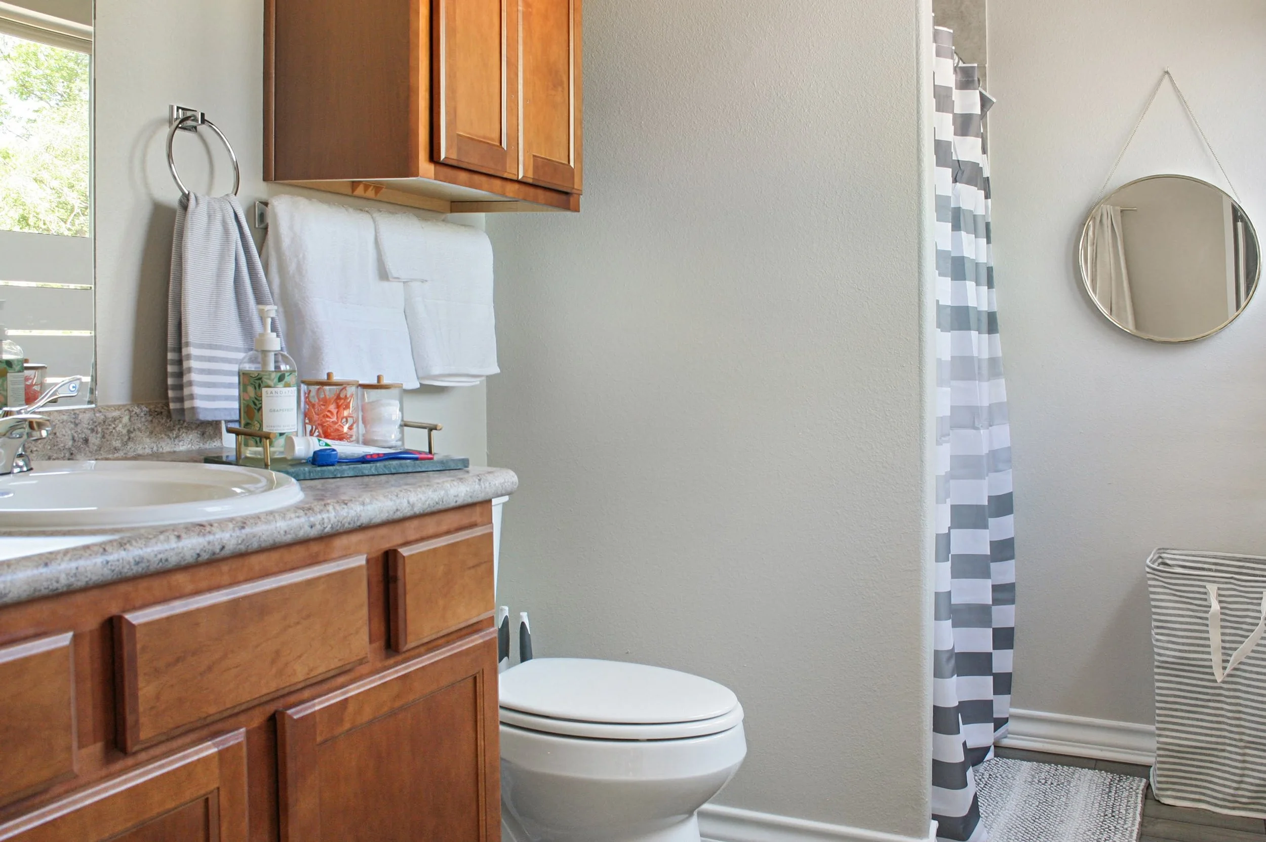 A bathroom with a vanity and sink, a mirror, towels, a shower with a striped curtain, and a laundry basket.