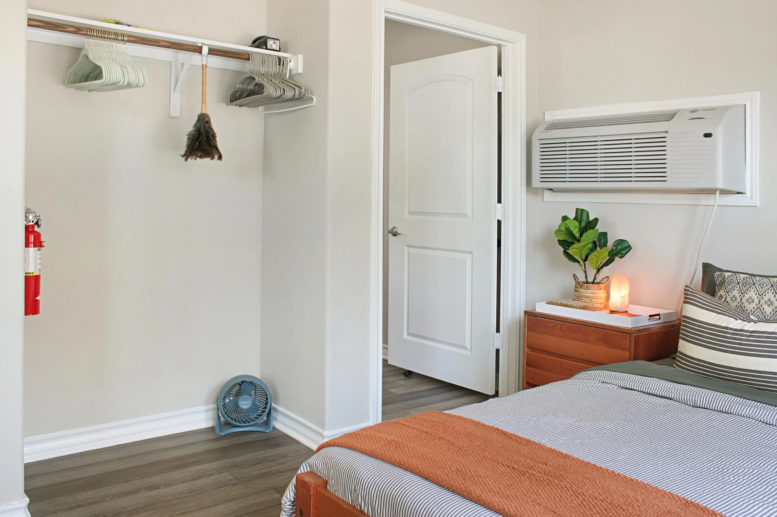 A bedroom with a bed, a nightstand with a plant, salt lamp, and books, a window AC unit, a closet with hangers and a small fan on the floor, and an open door.