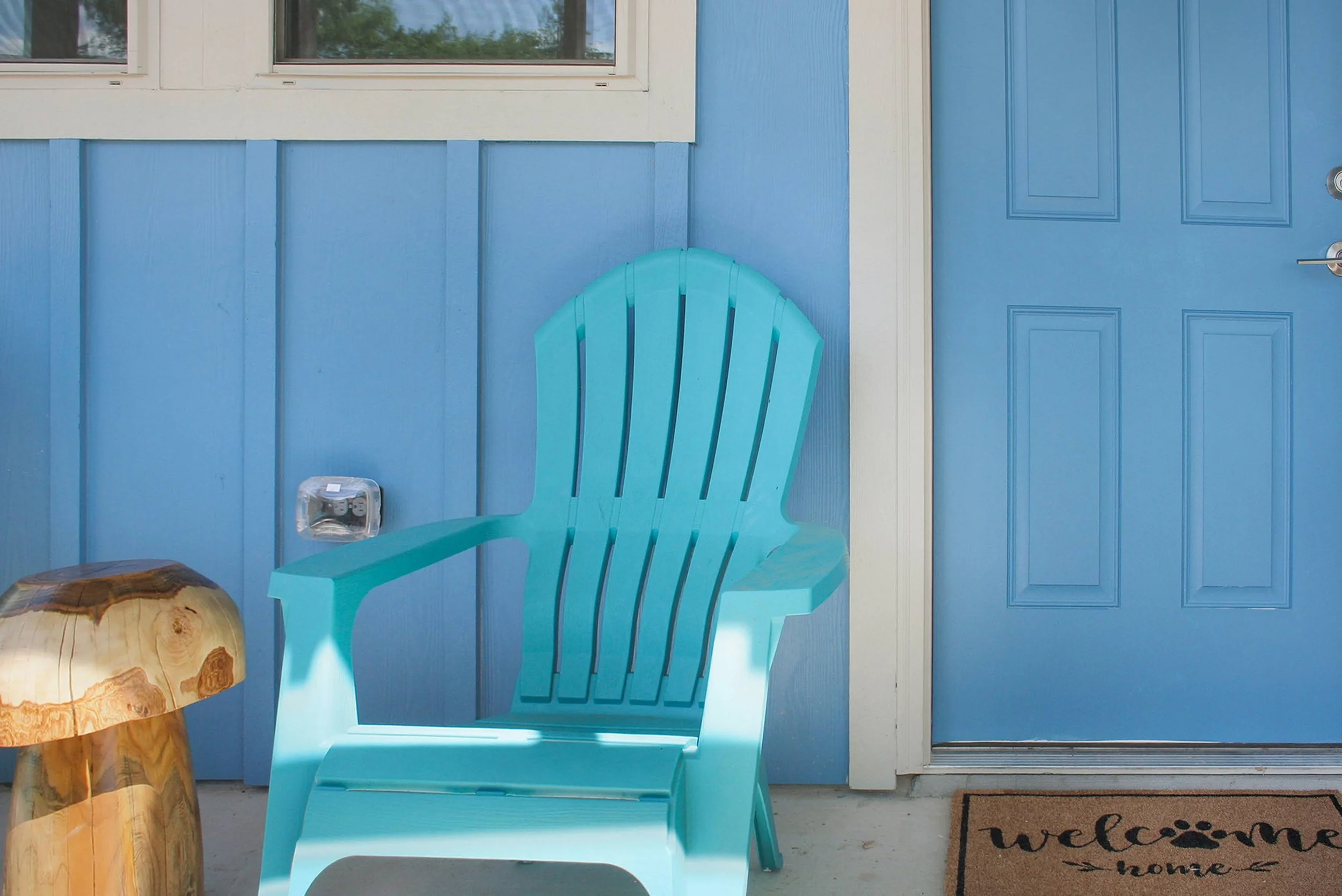 A porch with a blue wall, a door, a turquoise Adirondack chair, a small wooden side table, a window, and a welcome mat that says "welcome home."
