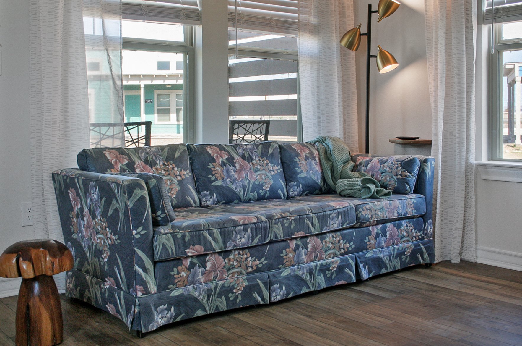 A vintage floral patterned sofa with green and pink flowers on a dark blue background in a bright living room with large windows, white curtains, a floor lamp with three brass shades, and a small wooden side table.