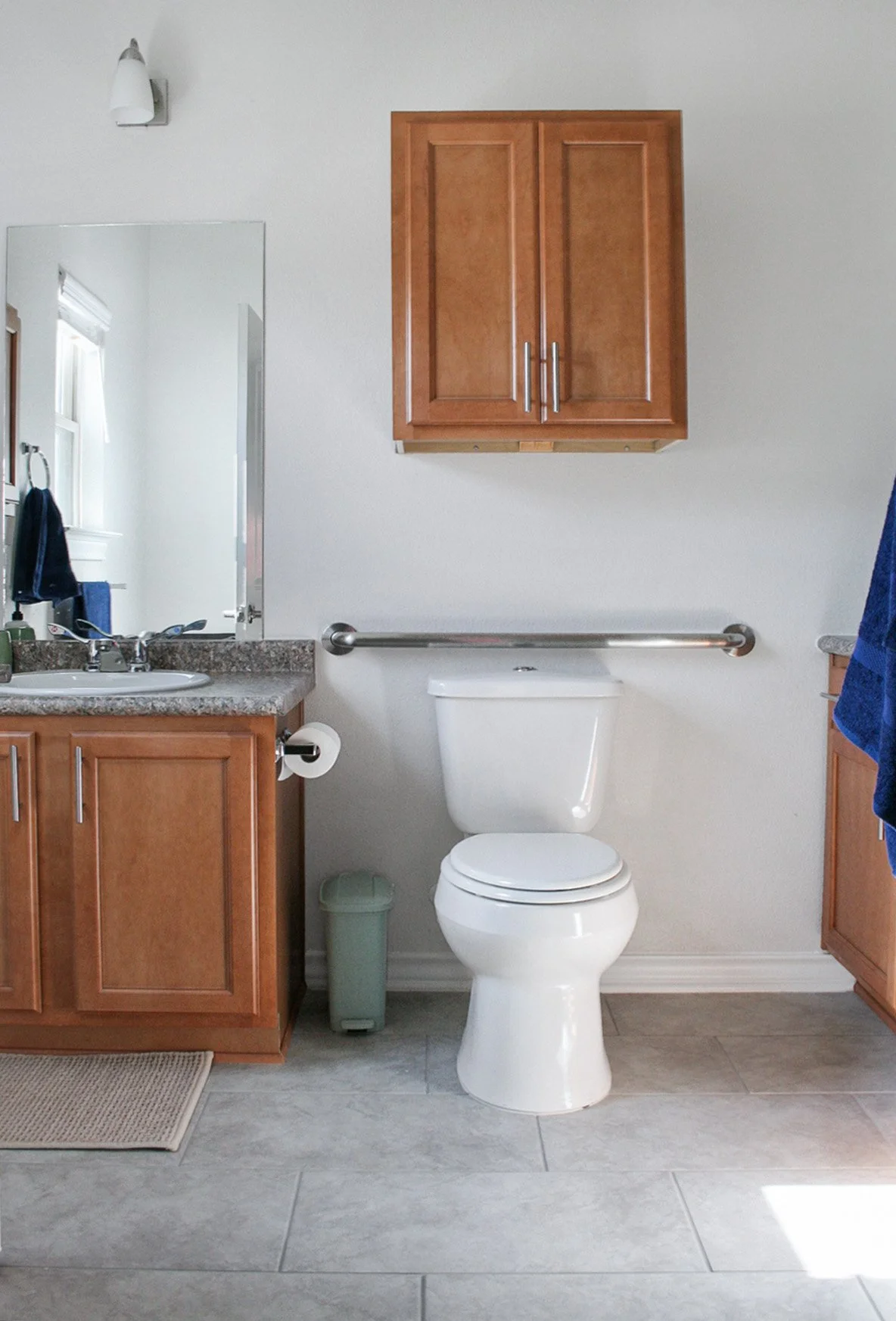 A bathroom with a white toilet, wooden cabinets, granite countertop sink, mirror, and a small green trash can.