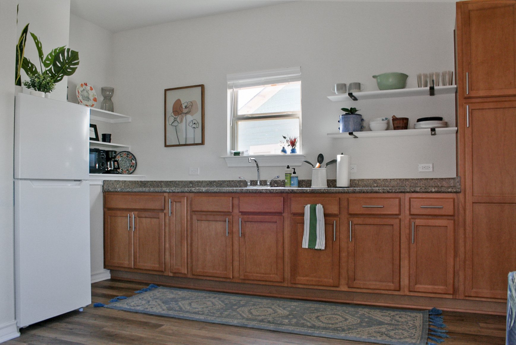 A kitchen with wooden cabinets, a granite countertop, a white refrigerator, open shelves with dishes and decor, a window above the sink with a small potted plant and decorative items, a patterned rug on a wooden floor, and a framed art piece on the w