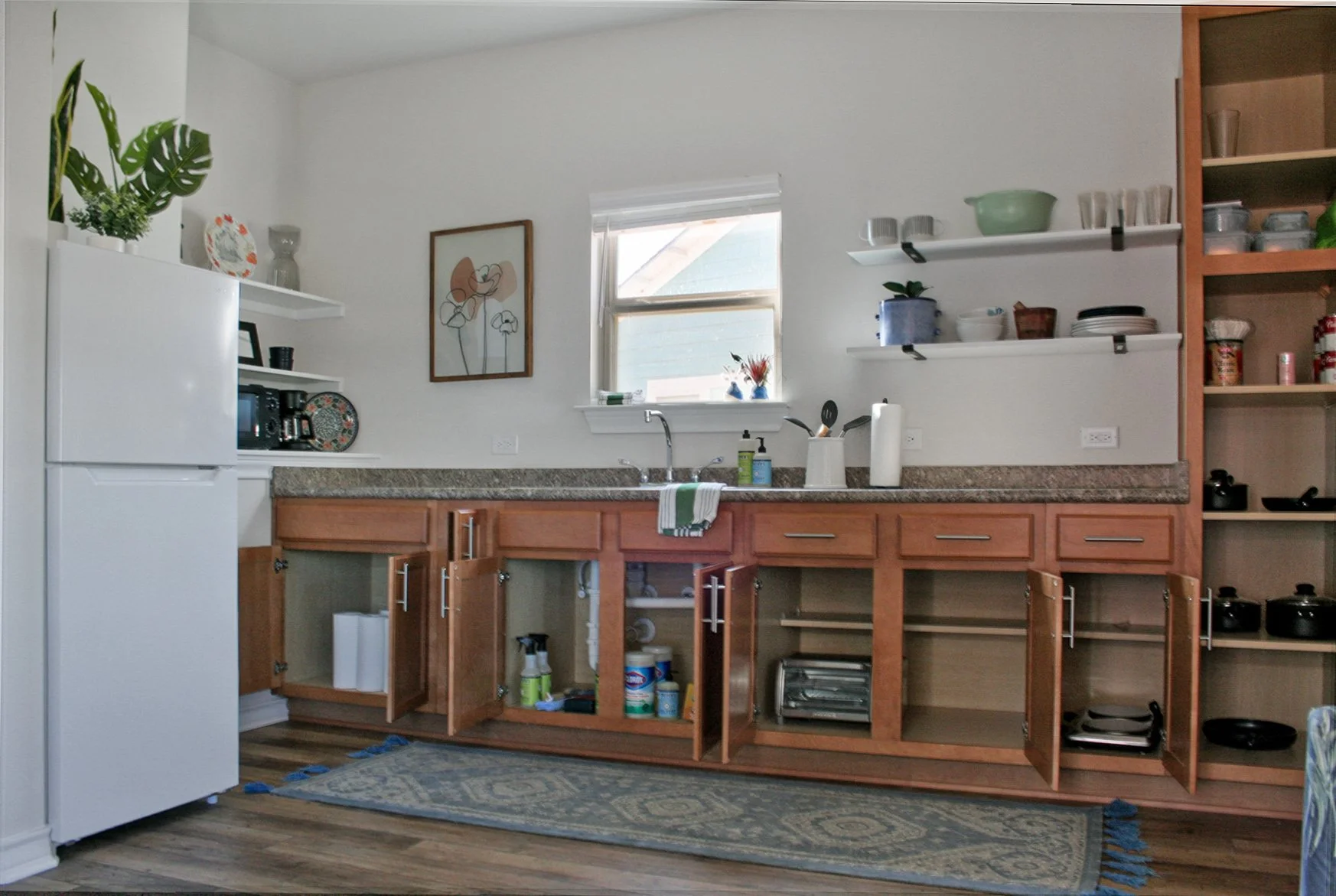 Kitchen with an open cabinet below the countertop, open shelves, and a white refrigerator on the left. The countertop has a sink with a window above, and various kitchen items. There is a framed picture on the wall, and a patterned rug on wooden floo