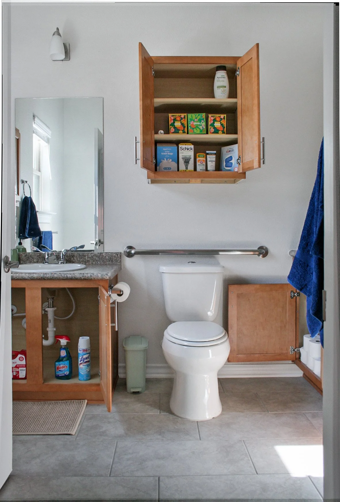 Small bathroom with a white toilet, a wooden cabinet above, and a countertop with a sink on the left, with cleaning supplies below and a mirror on the wall above.