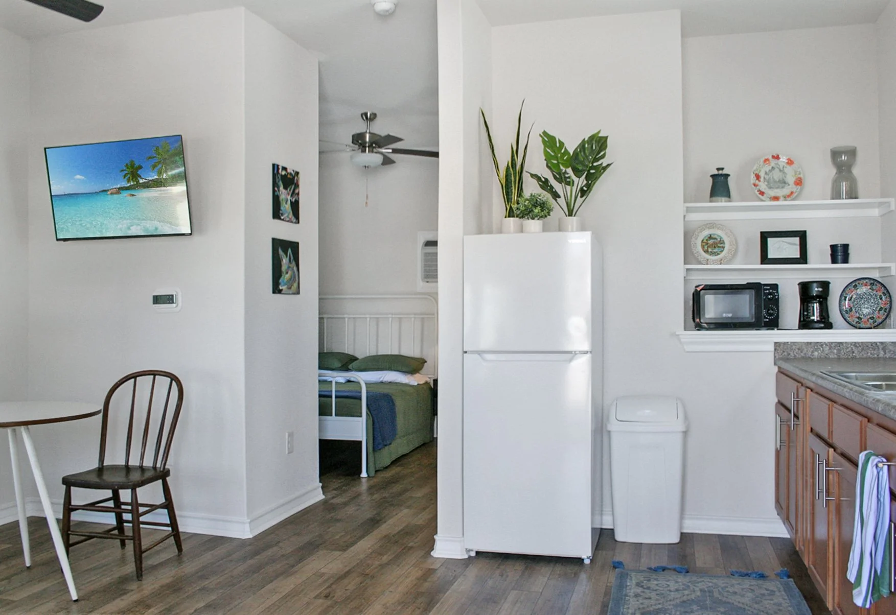 Interior of a small apartment with a kitchenette, a white refrigerator, and a mere glimpse of a bedroom with a bed and pillows in the background. There is a wall-mounted TV displaying a tropical beach scene, a small dining table with a wooden chair, 