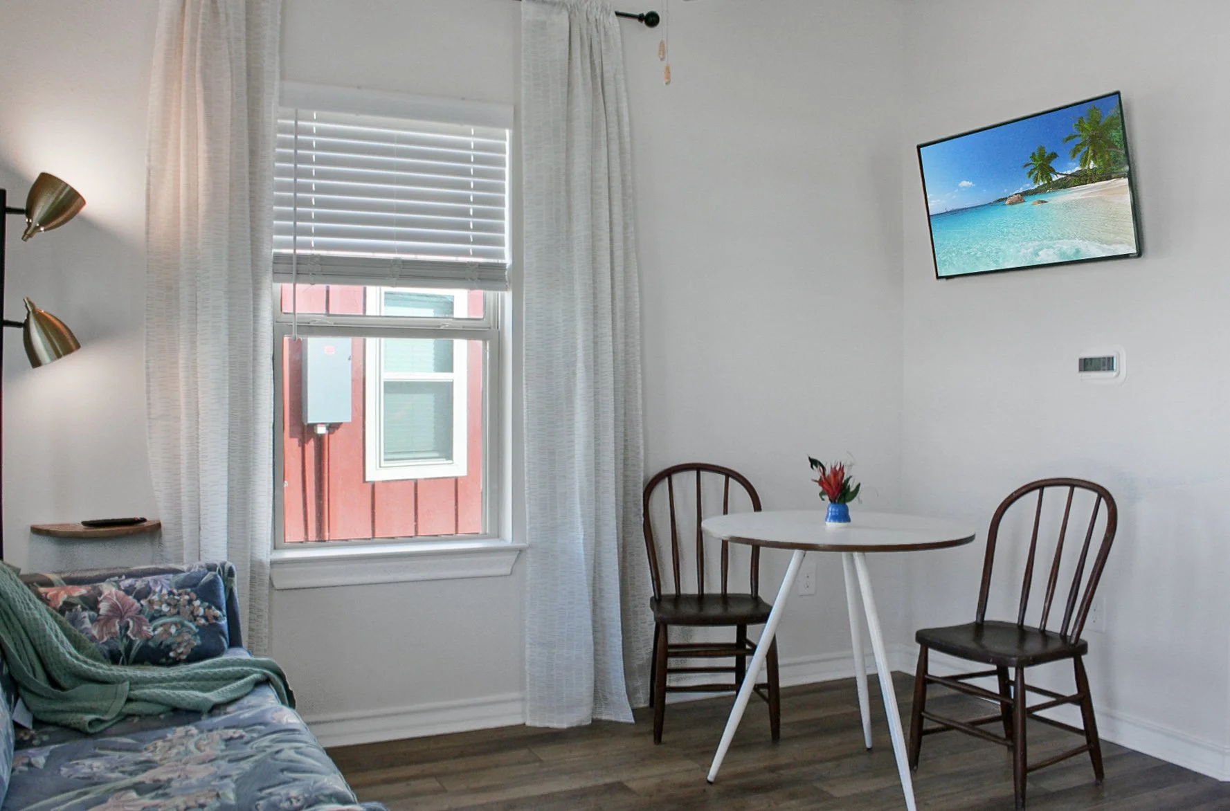 Interior of a cozy living space with a window, white curtains, a colorful floral couch, a small round table with two wooden chairs, a small vase with flowers, and a wall-mounted flat-screen TV showing a beach scene.