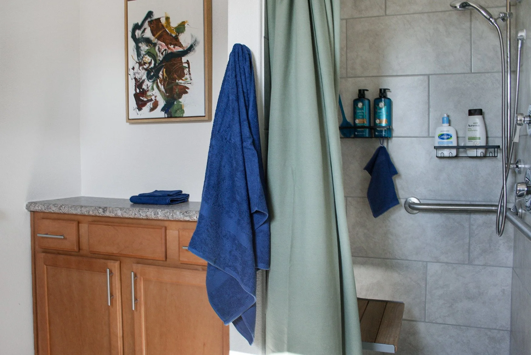 Bathroom with wooden cabinet, folded blue towels on top, colorful abstract painting on the wall, blue and green towels hanging, shower area with wall-mounted shelves holding shampoo, body wash, and lotion, handheld showerhead, and a green shower curt