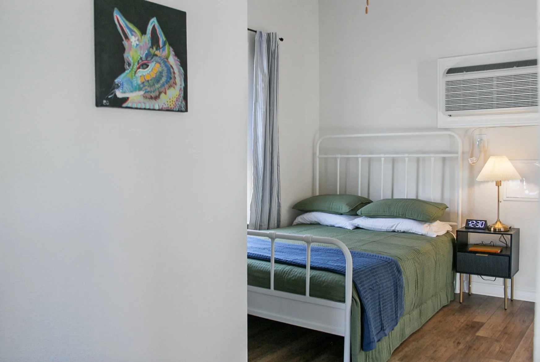 A bedroom with a bed that has green bedding and pillows, a small black nightstand with a digital clock displaying 12:30, a lamp, and an air conditioning unit, along with a framed colorful wolf artwork on the wall and a window with striped curtains.