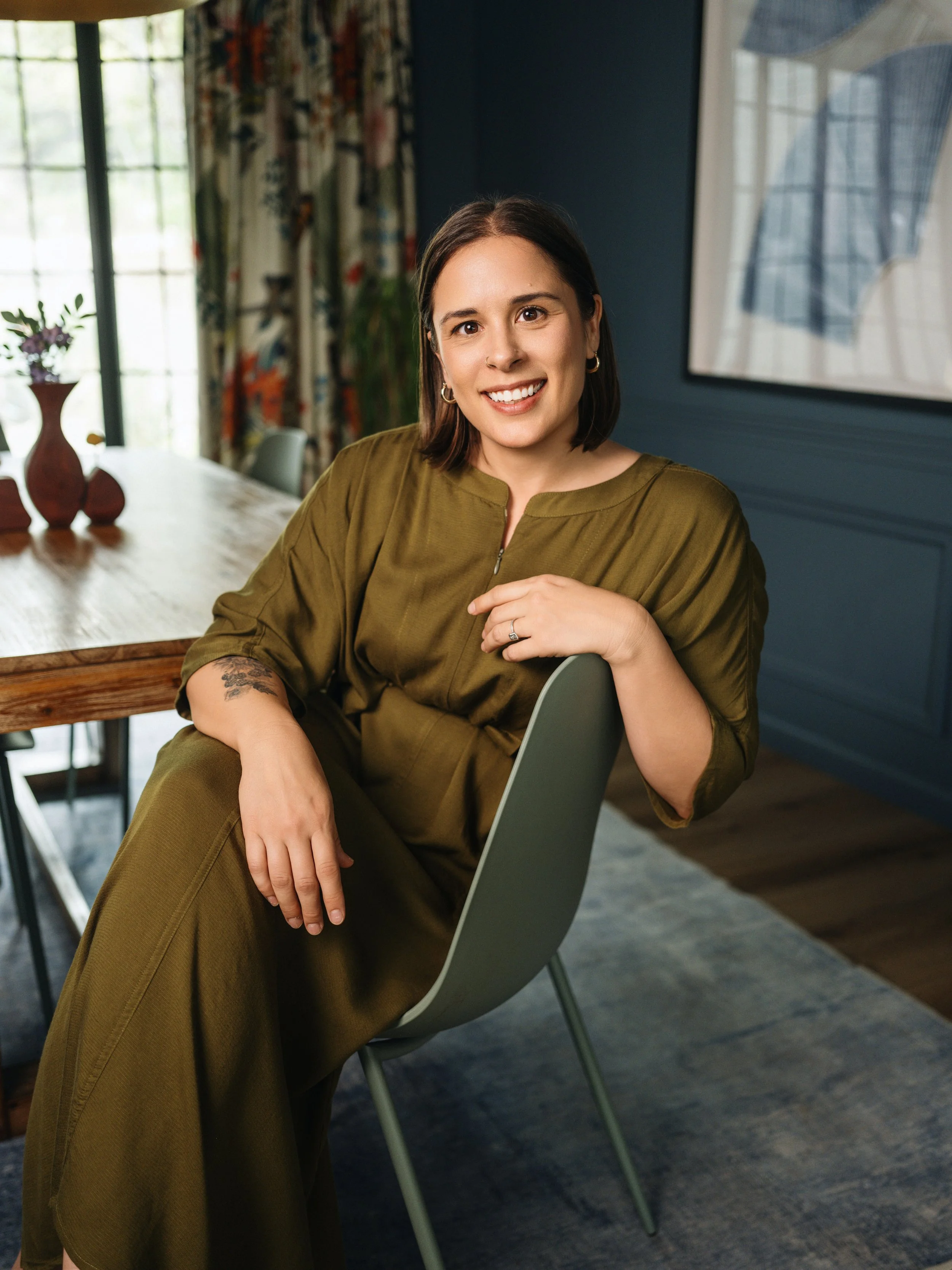 Woman with shoulder-length dark hair, wearing a green dress, sitting in a contemporary dining room with a big window, a framed artwork and a table with vases. She is smiling at the camera.