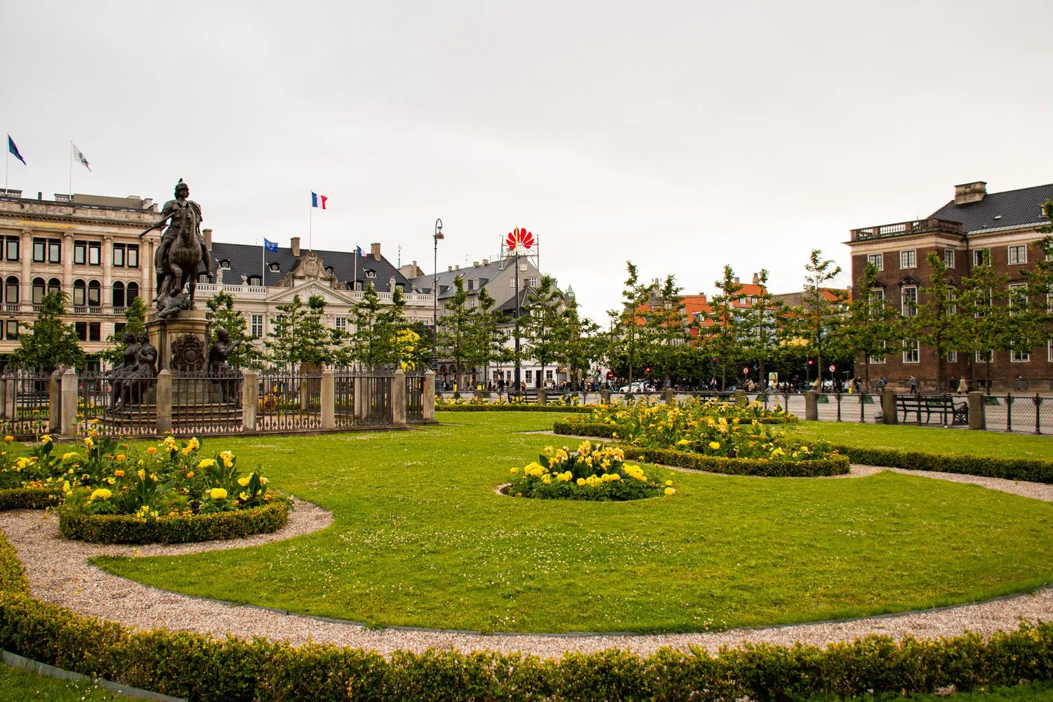 A picture of King’s Square on a cloudy summer day in Copenhagen. Flags blowing in the wind and yellow flowers in bloom. Also shown, a statue in the square center.