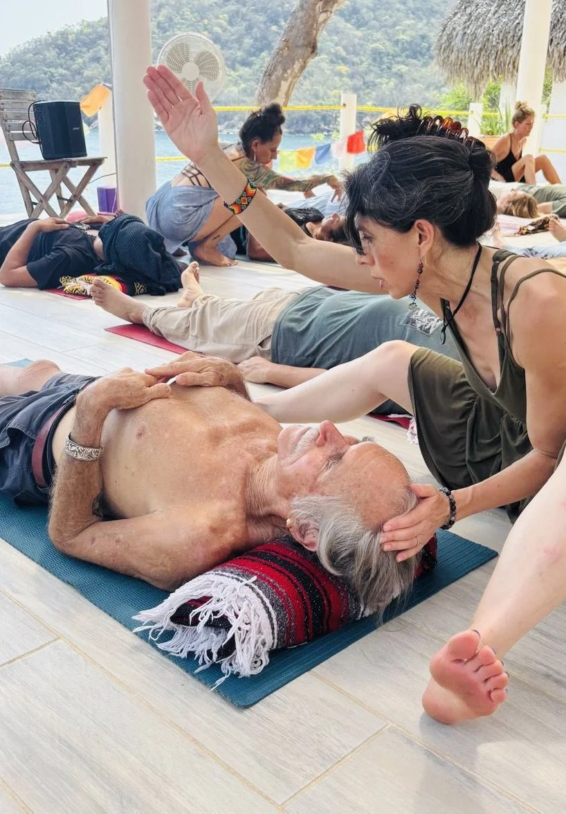 People participating in a yoga class on a covered outdoor deck overlooking water and trees, with an elderly man lying on a yoga mat receiving a gentle head massage from a woman.