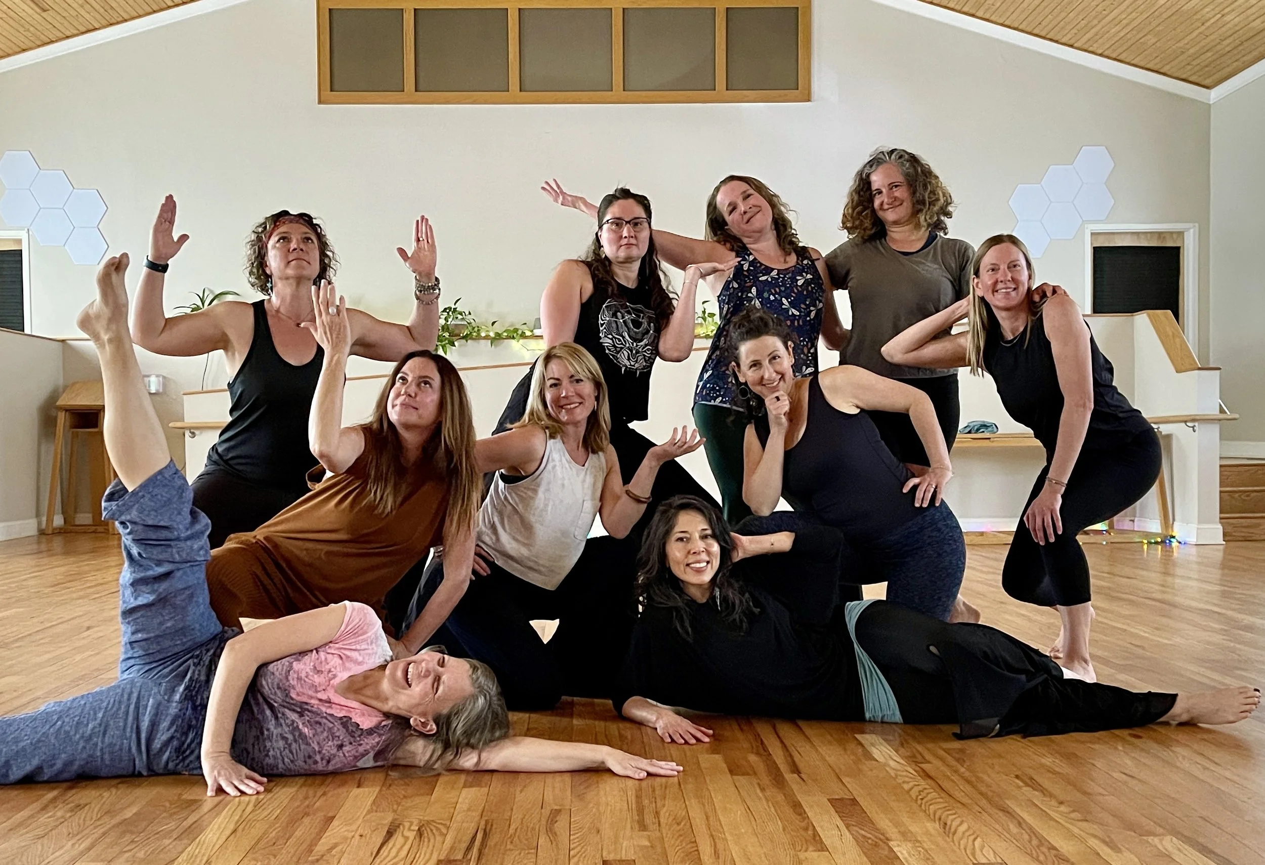 A group of ten women in a dance or yoga studio, posing playfully for the camera. Some women are standing, others are kneeling or lying on the wooden floor. The studio has light-colored walls, decorative hexagons, and some greenery.