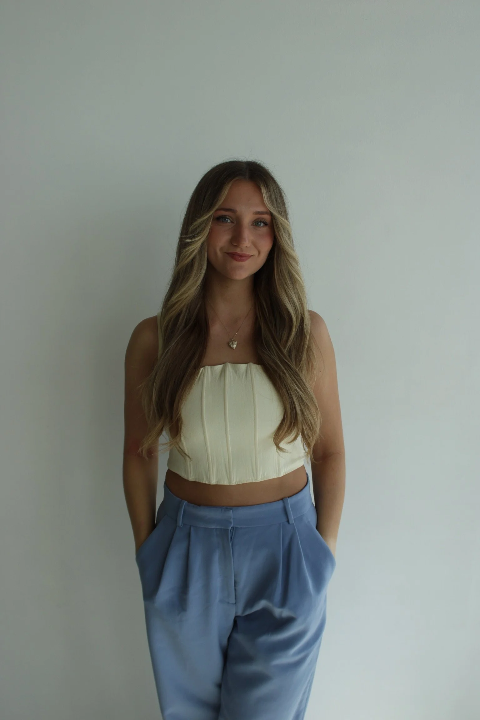 A woman with long wavy brown hair and blue eyes sitting on a white couch, wearing a brown blazer, black top, and white pants, smiling softly at the camera.