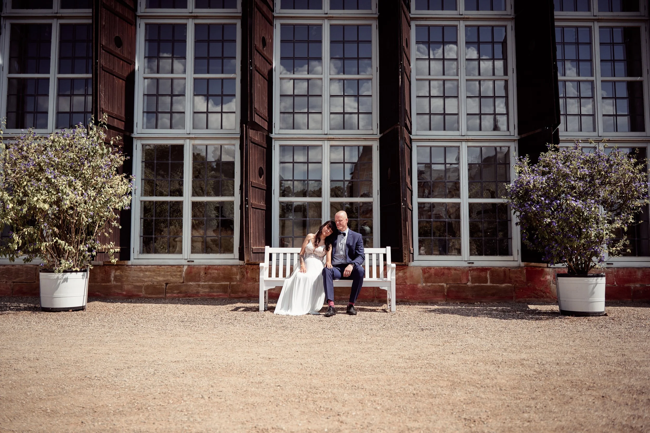 Ein Brautpaar sitzt auf einer weißen Bank vor einem Gebäude mit großen Fenstern und offenen Fensterläden. Links und rechts stehen große Blumentöpfe mit blühenden Sträuchern. Das Paar ist in formeller Hochzeitkleidung, das Mädchen trägt ein weißes Bra
