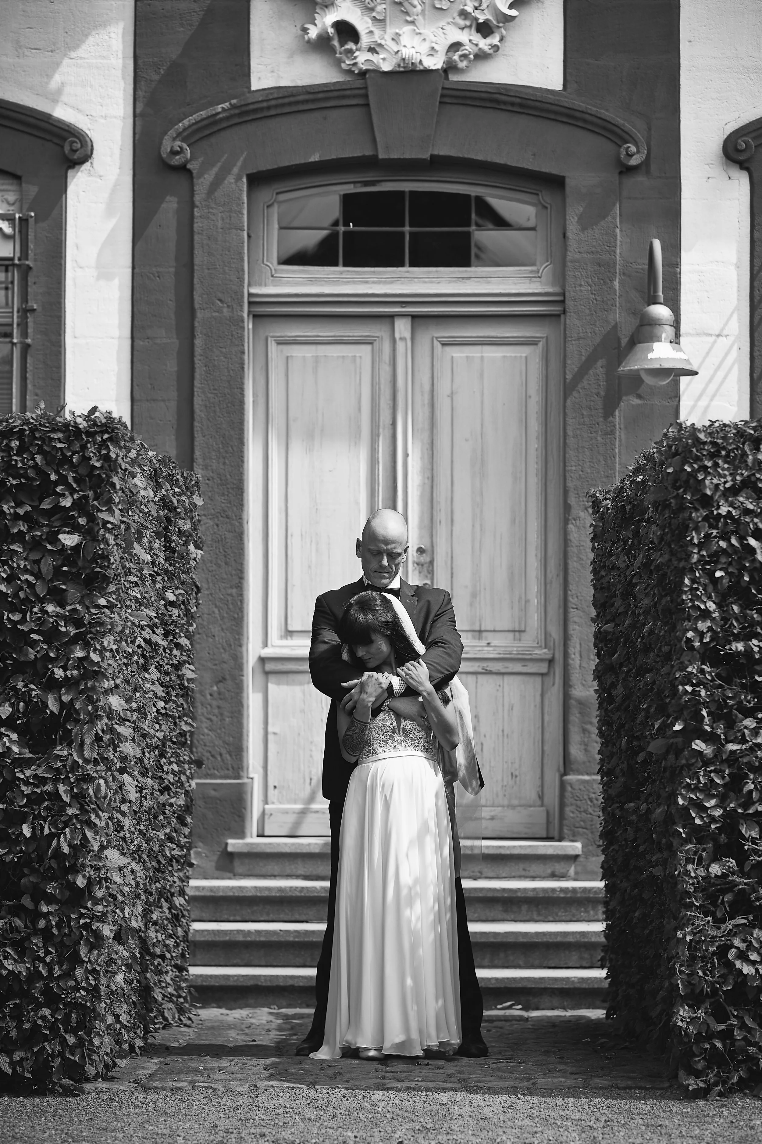 Ein Mann in einem Anzug hält eine Frau im Brautkleid schützend umarmt vor einer alten Tür.