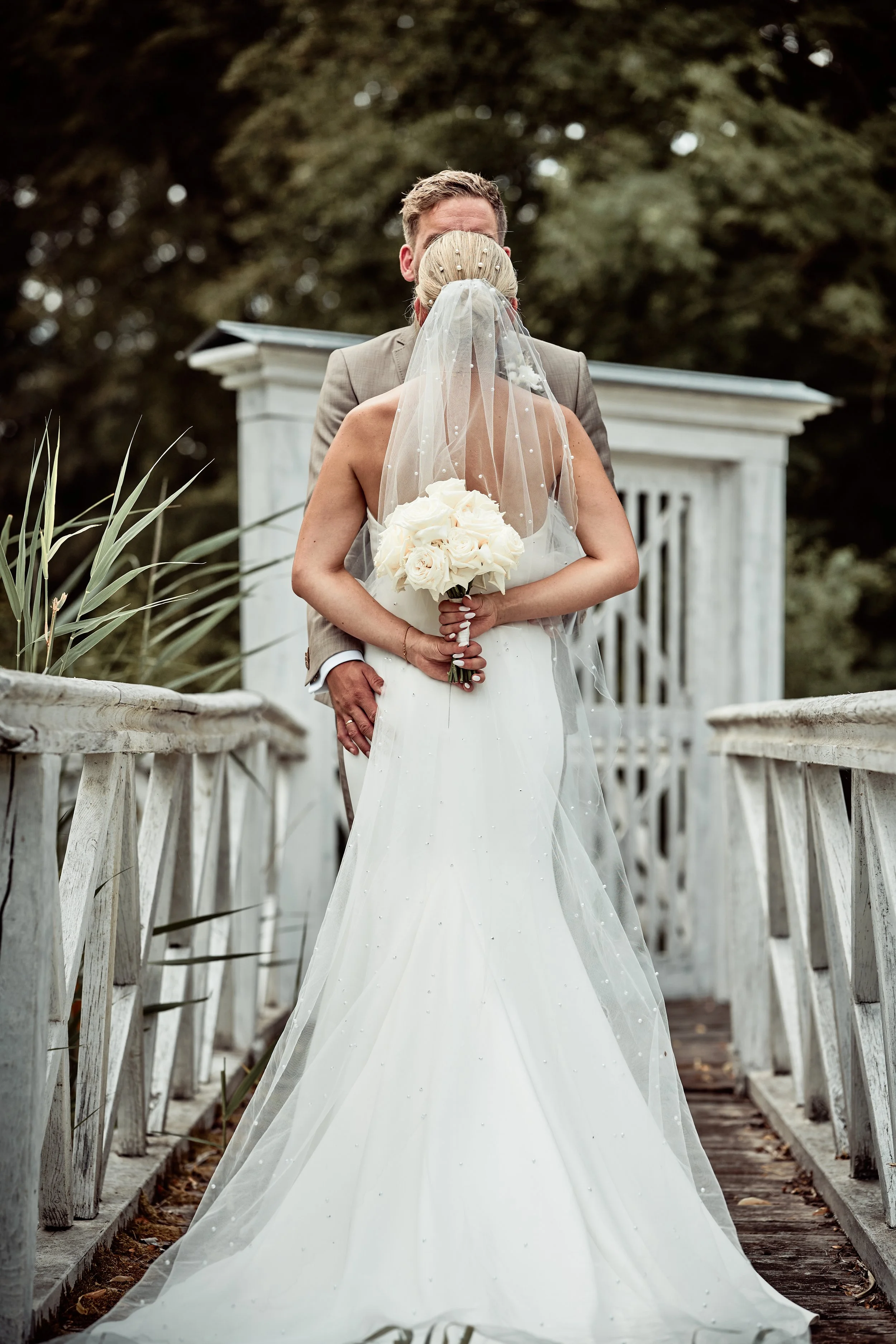 Braut und Bräutigam küssen sich auf einer Brücke bei der Hochzeit, Braut trägt ein weißes Kleid mit Schleier und hält einen Blumenstrauß, Bräutigam trägt einen Anzug.